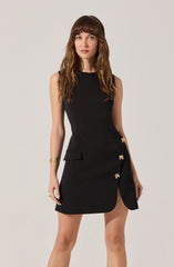 A woman is wearing a sleeveless, fitted black dress with a high neckline. The dress features gold embellishments on the front, including decorative clasps and bow-shaped accents, and has a front slit revealing part of her leg. She accessorized with gold jewelry, including a bracelet on her left wrist and rings on her fingers.