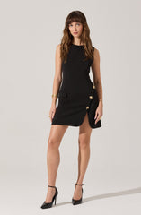 The woman is wearing a sleeveless black dress with a high neckline. The dress features decorative gold butterfly embellishments along the side, near a small slit that reveals part of her leg. She is also wearing black high-heeled shoes with ankle straps, and has gold jewelry on her left wrist and fingers.