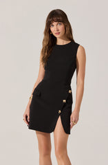 A young woman is wearing a sleeveless black dress with a fitted bodice and a skirt that features a high slit on one side. The dress is accented with decorative gold bow-shaped embellishments along the slit. She has long, wavy brown hair and is accessorized with gold jewelry, including a bracelet and rings.