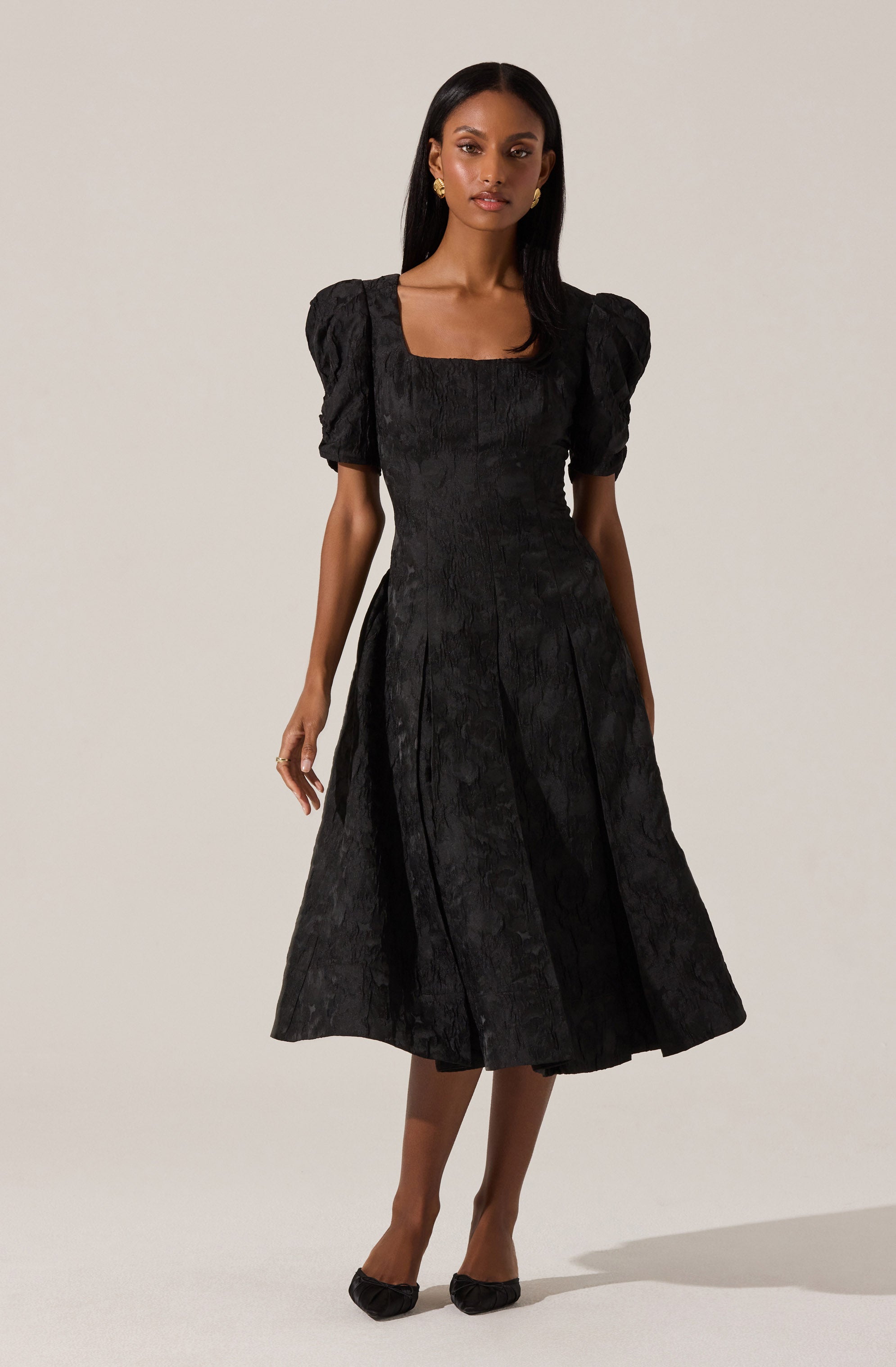 A woman is wearing a black, knee-length dress with a textured, floral pattern. The dress has puffed short sleeves, a square neckline, and pleats that create a flowing silhouette. She is also wearing black pointed-toe shoes and gold earrings.