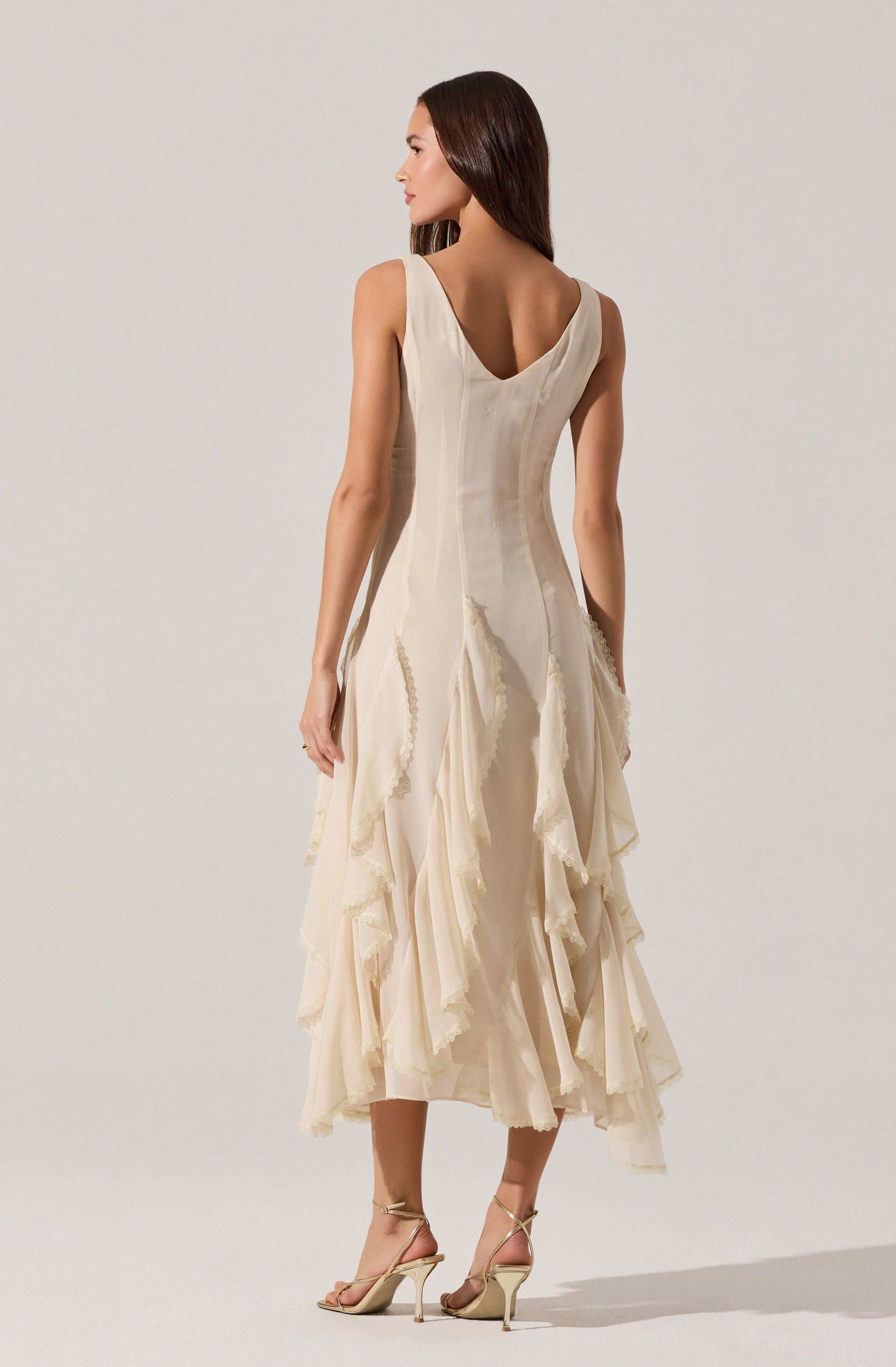 A woman is wearing an elegant sleeveless dress with a fitted bodice and a deep V neckline at the back. The dress is made of a light, cream-colored fabric with cascading, layered ruffles that run down from the waist to the hem, creating a textured, flowing appearance. The dress is midi-length with a slightly uneven hemline, and she is wearing strappy high-heeled sandals.