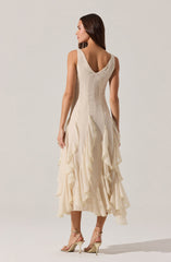 A woman is wearing an elegant sleeveless dress with a fitted bodice and a deep V neckline at the back. The dress is made of a light, cream-colored fabric with cascading, layered ruffles that run down from the waist to the hem, creating a textured, flowing appearance. The dress is midi-length with a slightly uneven hemline, and she is wearing strappy high-heeled sandals.