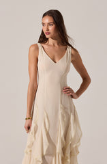 A woman is wearing an elegant sleeveless cream-colored dress with a V-neckline. The dress features vertical seam details on the bodice and layered, ruffled fabric accents with lace trim along the edges starting from the waist down to the hem, creating a flowing, textured look. She has minimal accessories, including a gold bracelet and ring, and her long brown hair is styled in loose waves.