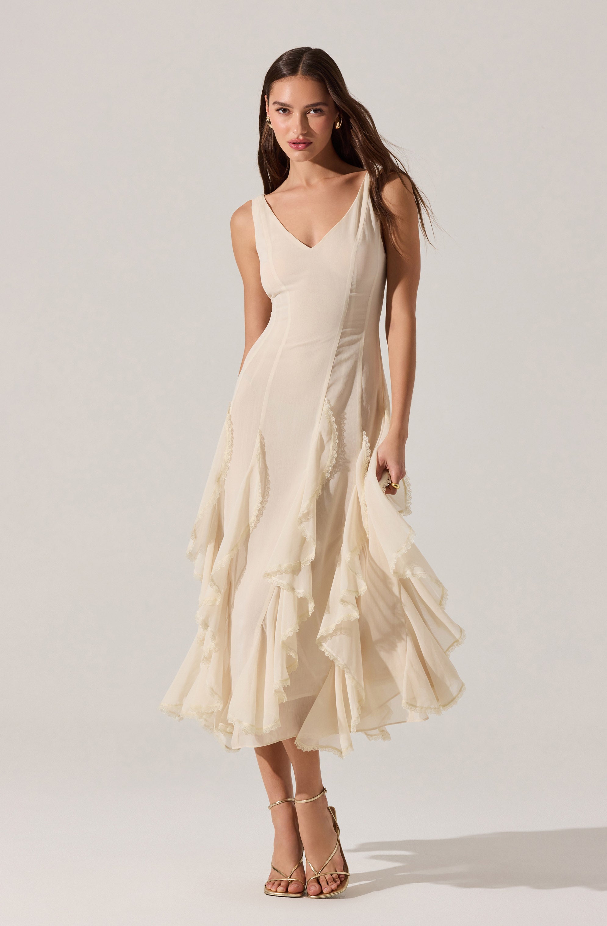 A woman is wearing a sleeveless, cream-colored dress with a V-neckline. The dress features layered, ruffled fabric panels that create a textured, flowing look. She has on strappy high-heeled sandals in a metallic or neutral tone.