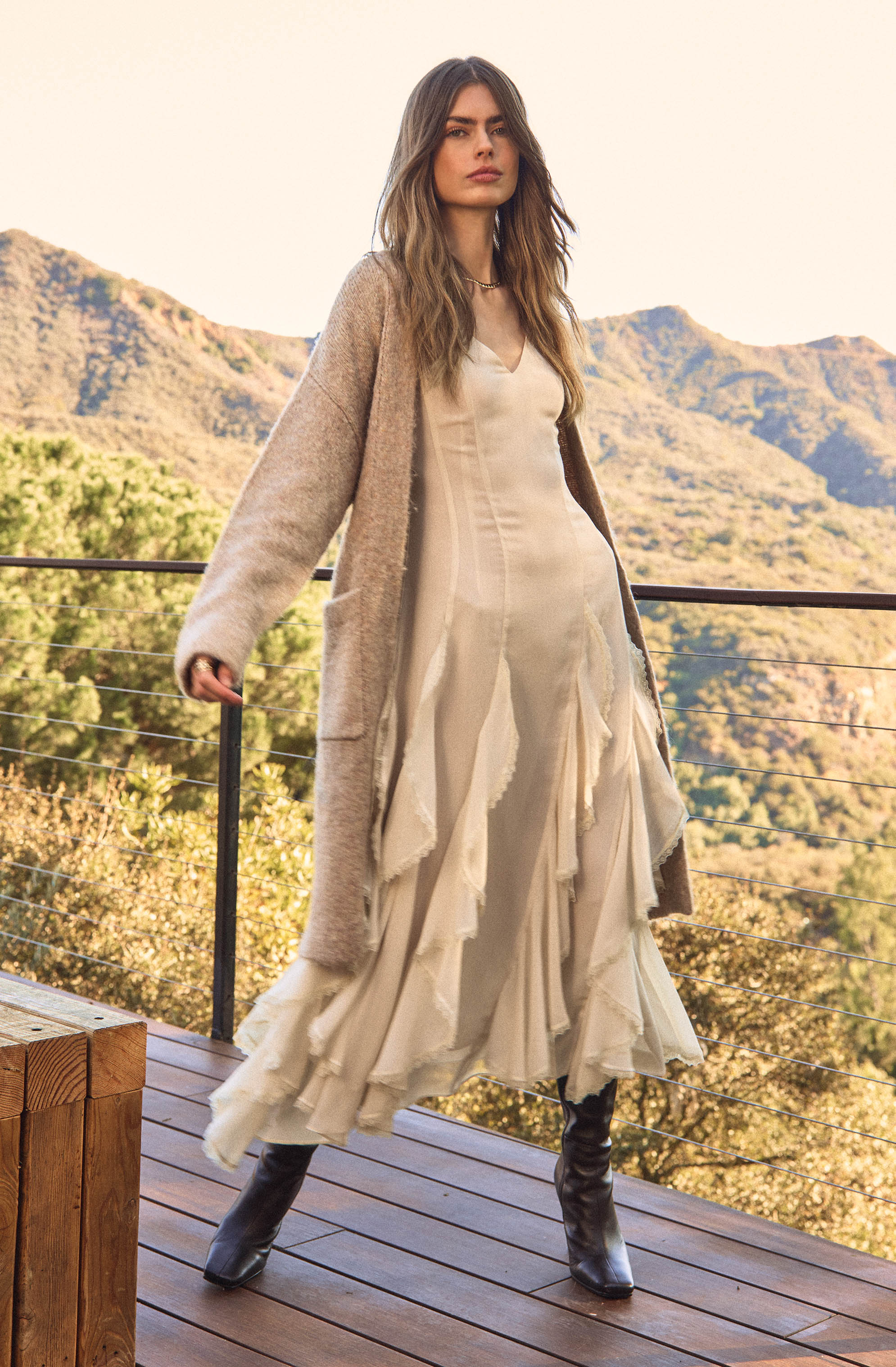 A woman is wearing a long, flowing cream-colored dress with layered, ruffled fabric details. She has on a beige, textured, open-front cardigan that extends to her knees. She completes her outfit with black knee-high boots.