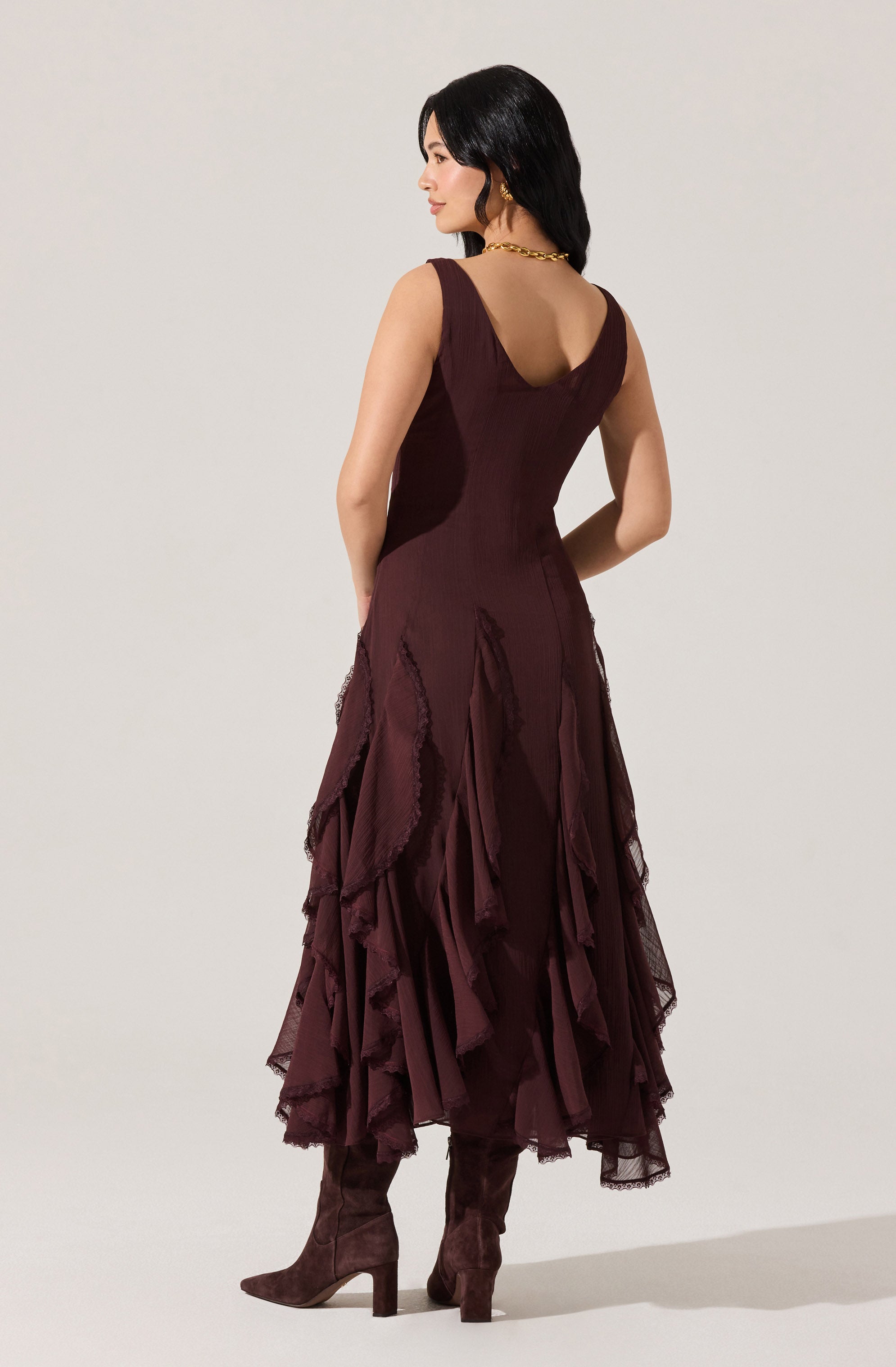 A woman is wearing a sleeveless, deep burgundy dress with a fitted bodice and a V-shaped neckline. The skirt is long and layered with ruffled, uneven panels made of sheer, textured fabric, creating a voluminous and dramatic look. She is also wearing matching high-heeled, knee-high boots in the same burgundy color.