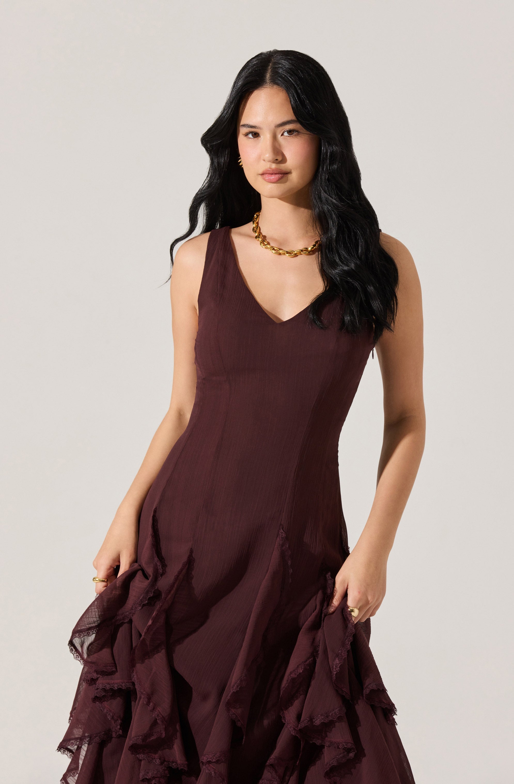 A woman is wearing a sleeveless, deep burgundy dress with a V-neckline. The dress has a fitted bodice and features layered ruffle details at the skirt. She accessorizes with a chunky gold chain necklace and gold hoop earrings.