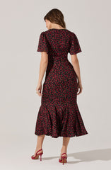 A woman is wearing a black dress with a red floral pattern. The dress has short, puffed sleeves and features a fitted waist with a gathered, ruffled hem at the bottom. She is also wearing red high-heeled shoes.