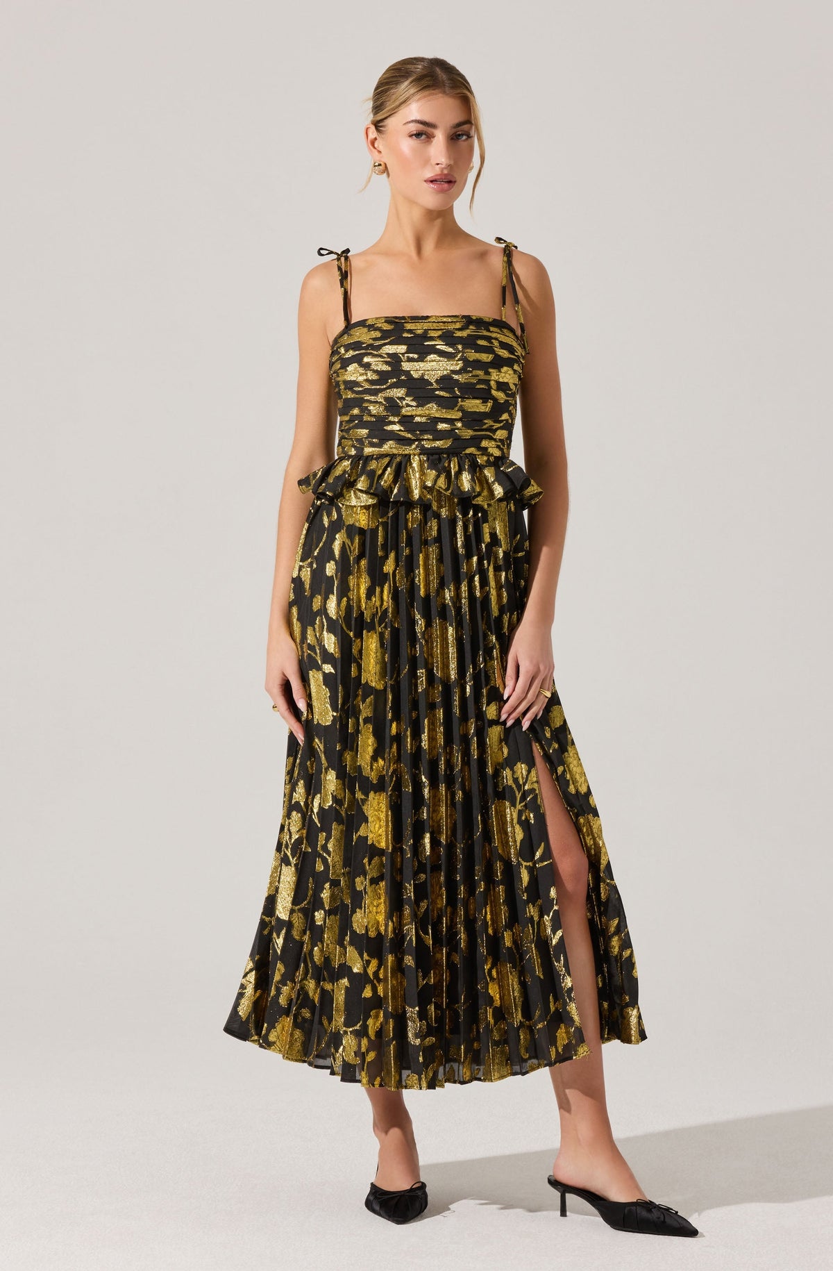 A woman is wearing a sleeveless, black and gold dress with a leopard print pattern. The dress has thin, tie-up shoulder straps, a gathered bodice, and a high slit on one side. The fabric is pleated, adding texture and movement to the skirt. She is wearing black heeled shoes.