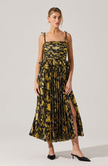A woman is wearing a sleeveless, black and gold dress with a leopard print pattern. The dress has thin, tie-up shoulder straps, a gathered bodice, and a high slit on one side. The fabric is pleated, adding texture and movement to the skirt. She is wearing black heeled shoes.