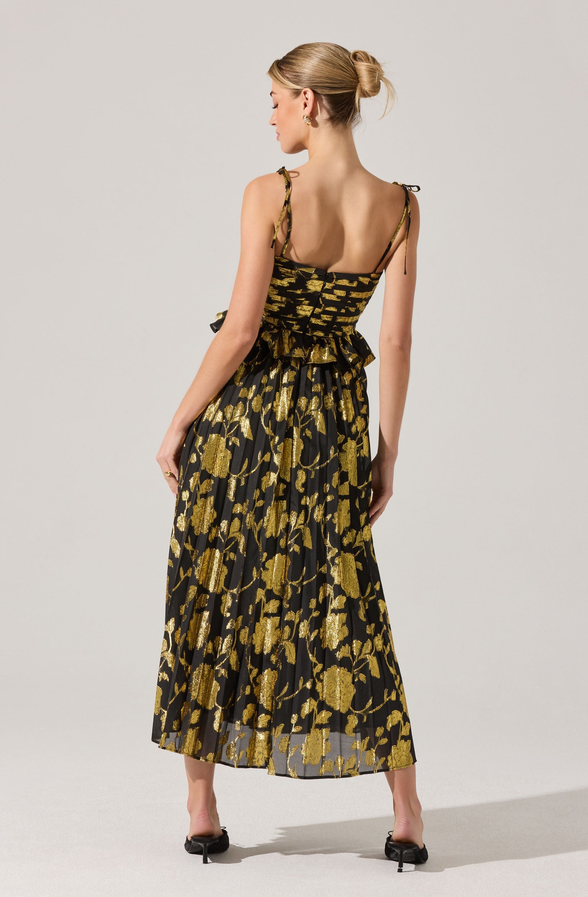 A woman is wearing a sleeveless dress with adjustable shoulder ties. The dress features a fitted bodice with a flared, ruffled waist. The fabric has a black base with a gold floral pattern throughout, and the skirt is long and flowing, reaching her ankles. She is also wearing black open-toe heels.