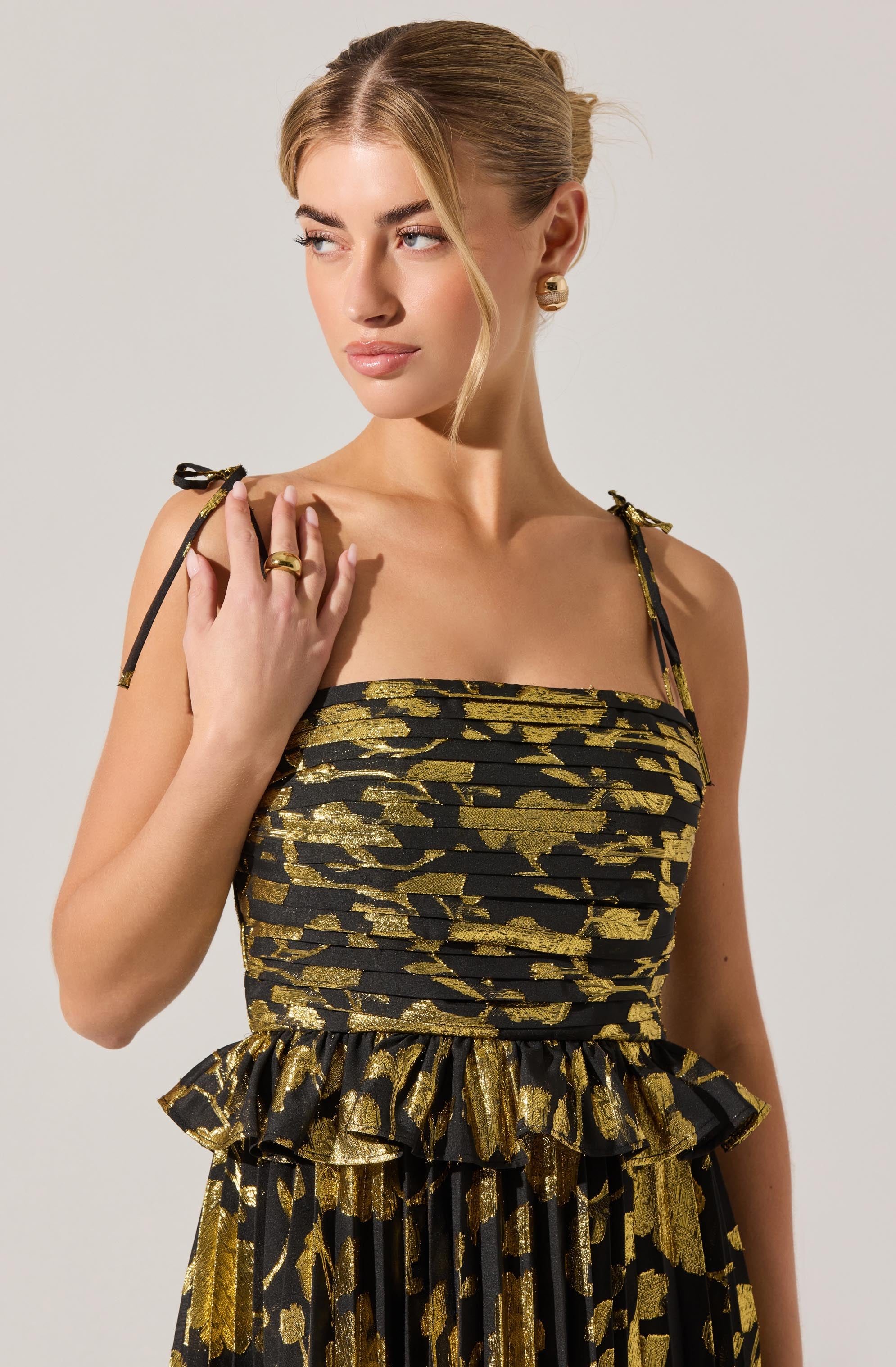 A woman is wearing a sleeveless dress with black and gold abstract patterns. The dress has thin, adjustable shoulder straps tied into small bows, and features a fitted bodice with a ruffled peplum detail at the waist.