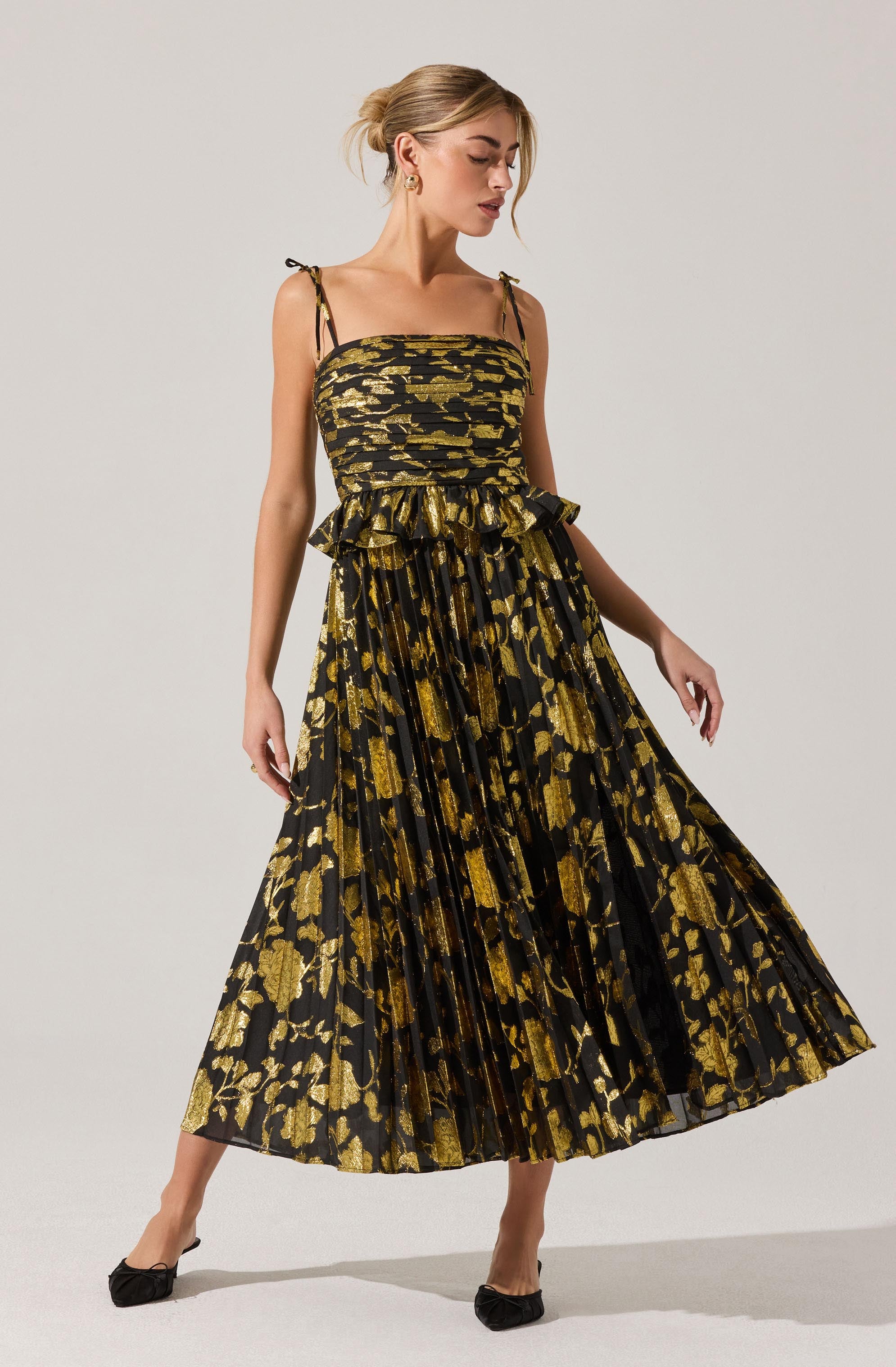 A woman is wearing a sleeveless, ankle-length dress with thin, adjustable shoulder straps tied into bows. The dress features a fitted bodice and a flared, pleated skirt. The fabric has a black background with a bold gold floral and leaf pattern throughout.
