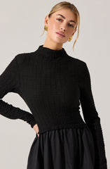 A woman is wearing a fitted, long-sleeved black textured sweater with a high, mock neckline. She pairs it with a black skirt that has a gathered, slightly shiny fabric. She accessorizes with large, round gold earrings featuring a spiral design.