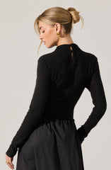 A woman is wearing a fitted, long-sleeved black sweater with a textured, chunky knit pattern. The sweater has a small opening at the back near the neckline, closed with a button. She pairs it with a high-waisted, black skirt made of a shiny, slightly puffy fabric. She accessorizes with large, gold earrings. Her hair is styled in a low, messy bun.