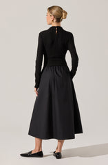 A woman is wearing a long black pleated skirt that reaches her mid-calf, paired with a textured black long-sleeve top with a keyhole opening at the back. She completes her outfit with black flat shoes.