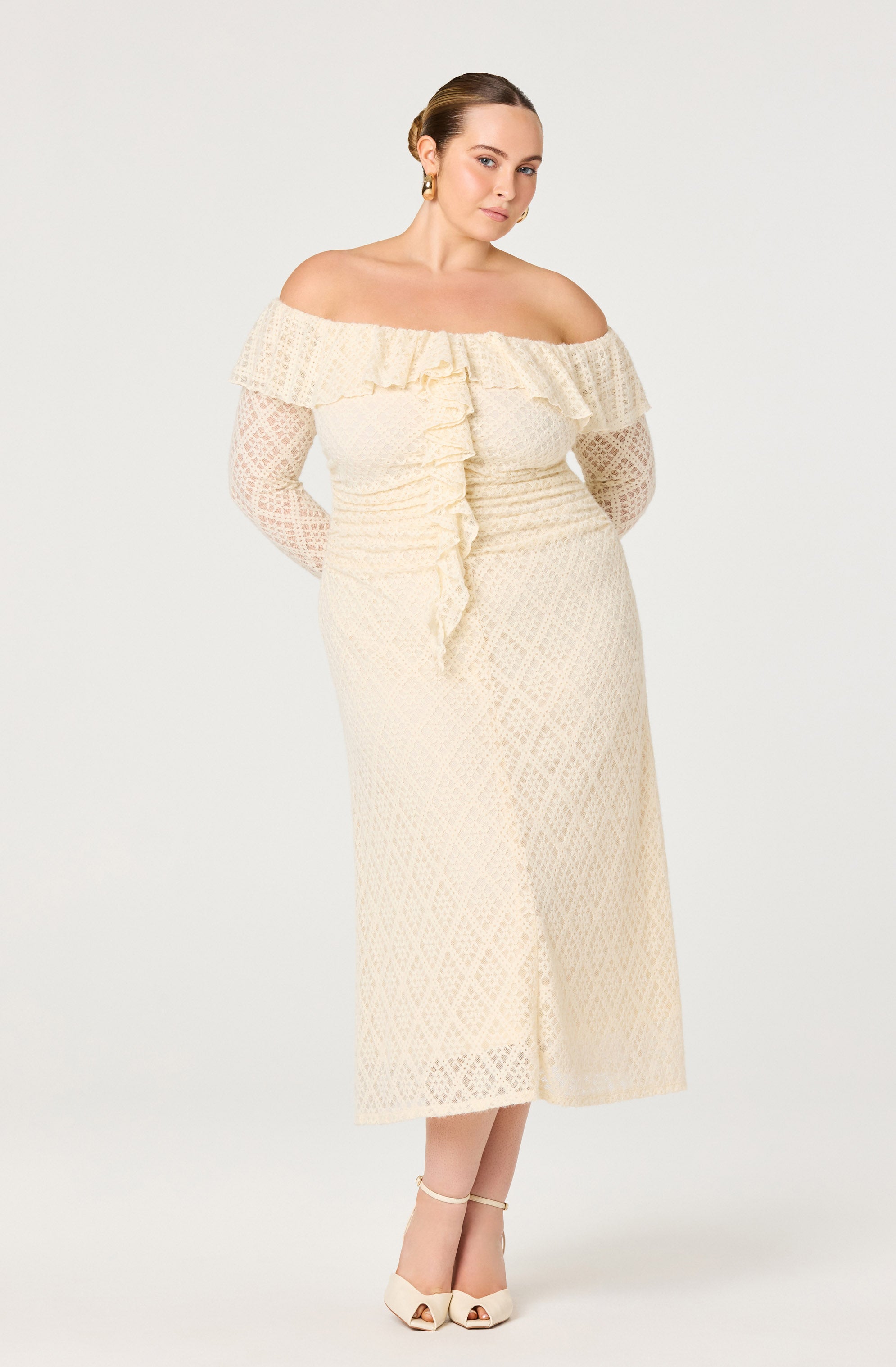 A woman is wearing an off-the-shoulder cream-colored dress with a ruffled neckline and lace long sleeves. The dress features a subtle diamond pattern, a front ruffle detail that runs vertically down the center, and a midi length. She is also wearing cream-colored high-heeled sandals with ankle straps.