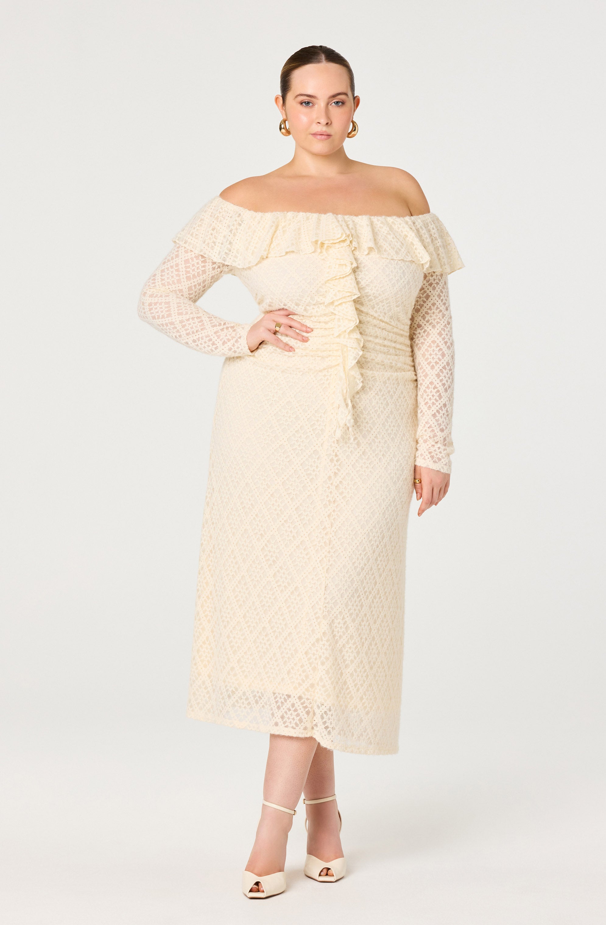 The woman is wearing a cream-colored, off-the-shoulder dress with long, semi-sheer lace sleeves. The dress features a ruffled neckline that extends around the shoulders and a front ruffle detail running down the center. She is also wearing cream-colored high-heeled shoes with ankle straps.