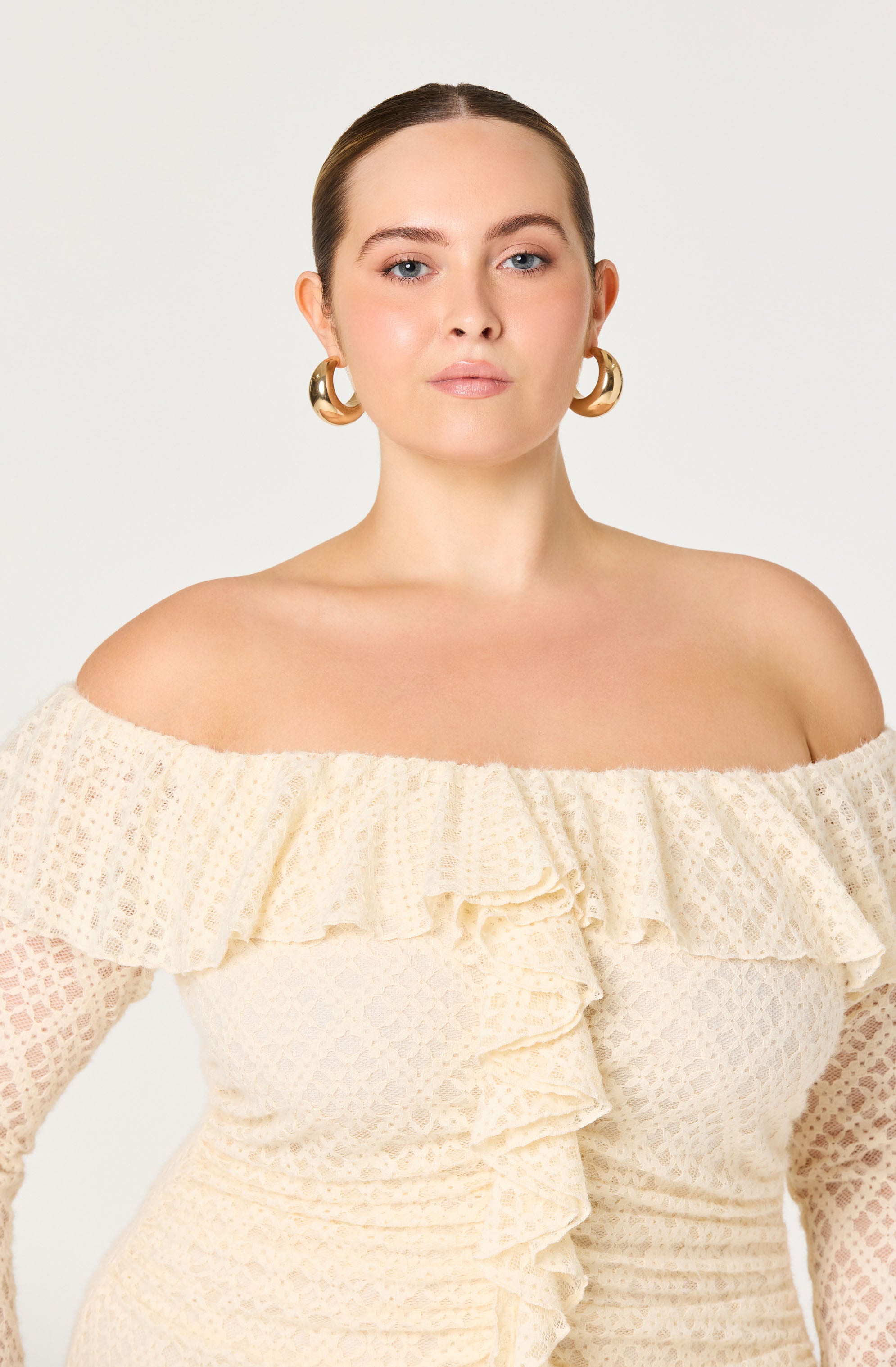 A woman is wearing an off-the-shoulder cream-colored dress with a textured, lace-like pattern. The dress features ruffles across the neckline and vertical ruffles down the front. She has large, gold hoop earrings and her hair is pulled back.