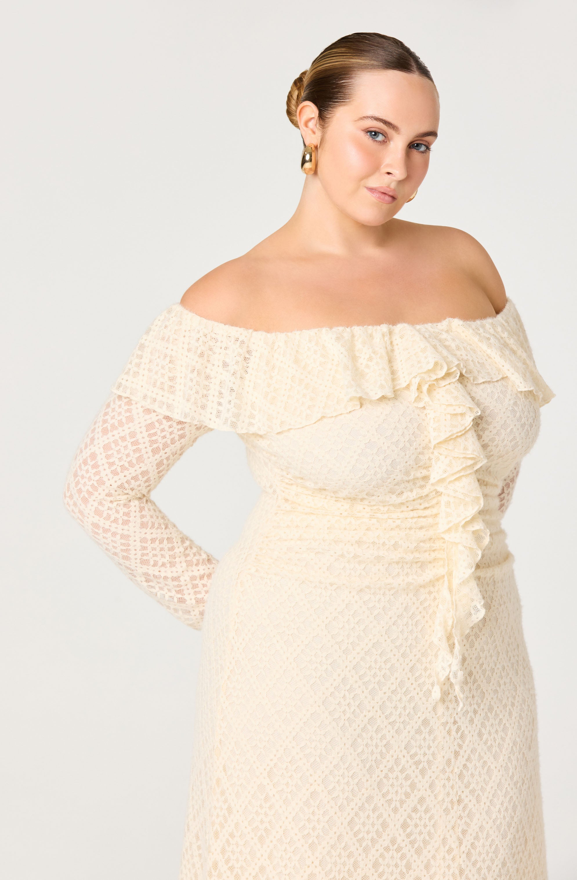 A woman is wearing an off-the-shoulder, long-sleeve dress made of cream-colored lace fabric. The dress features a ruffled neckline that extends across the shoulders and down the front, with a textured pattern throughout. The fabric has a delicate, openwork lace design, and the dress fits snugly around her waist.