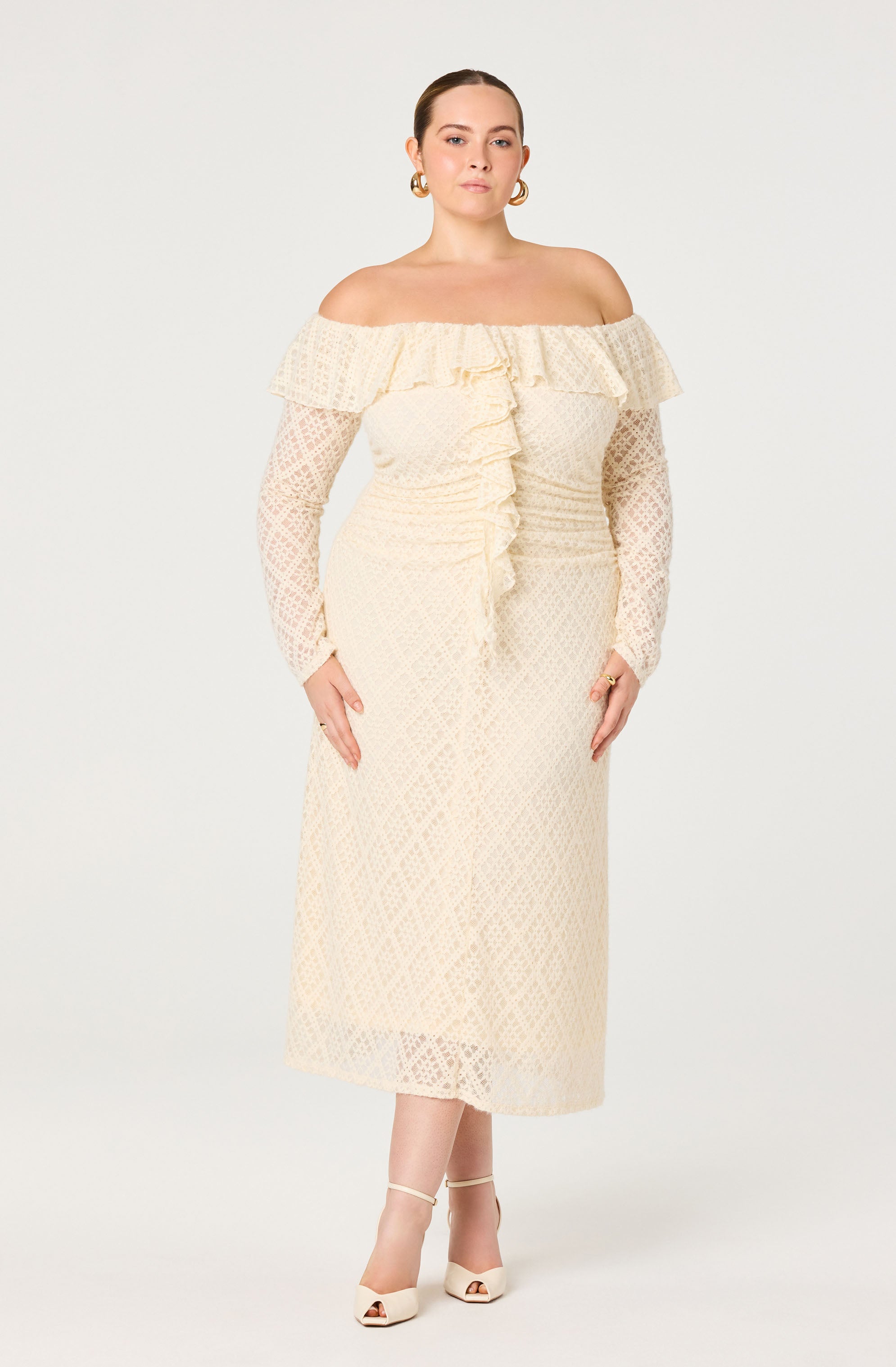 A woman is wearing an off-the-shoulder, long-sleeved, cream-colored dress with intricate lace details. The dress features a ruffled neckline and a ruffled front running vertically down the center. She is accessorized with gold hoop earrings and cream-colored open-toe heels with ankle straps.