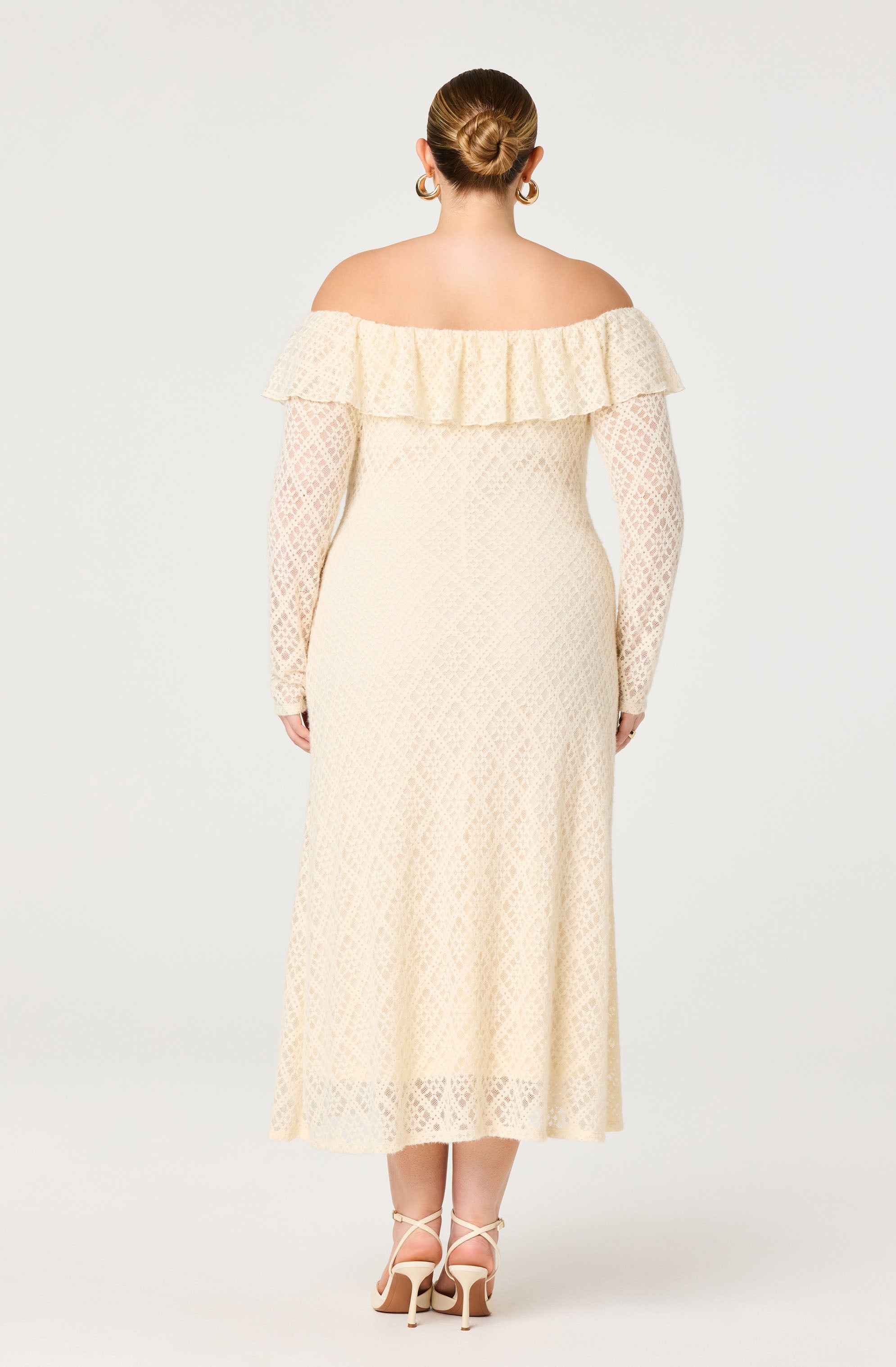 The dress is an off-the-shoulder design featuring a large ruffle across the neckline. It has long, fitted sleeves made of lace with a floral pattern. The fabric of the dress has a delicate, diamond-shaped lace pattern throughout, creating a semi-sheer look that extends to a midi length, ending just below the calves. The dress is in a pale cream or ivory color.