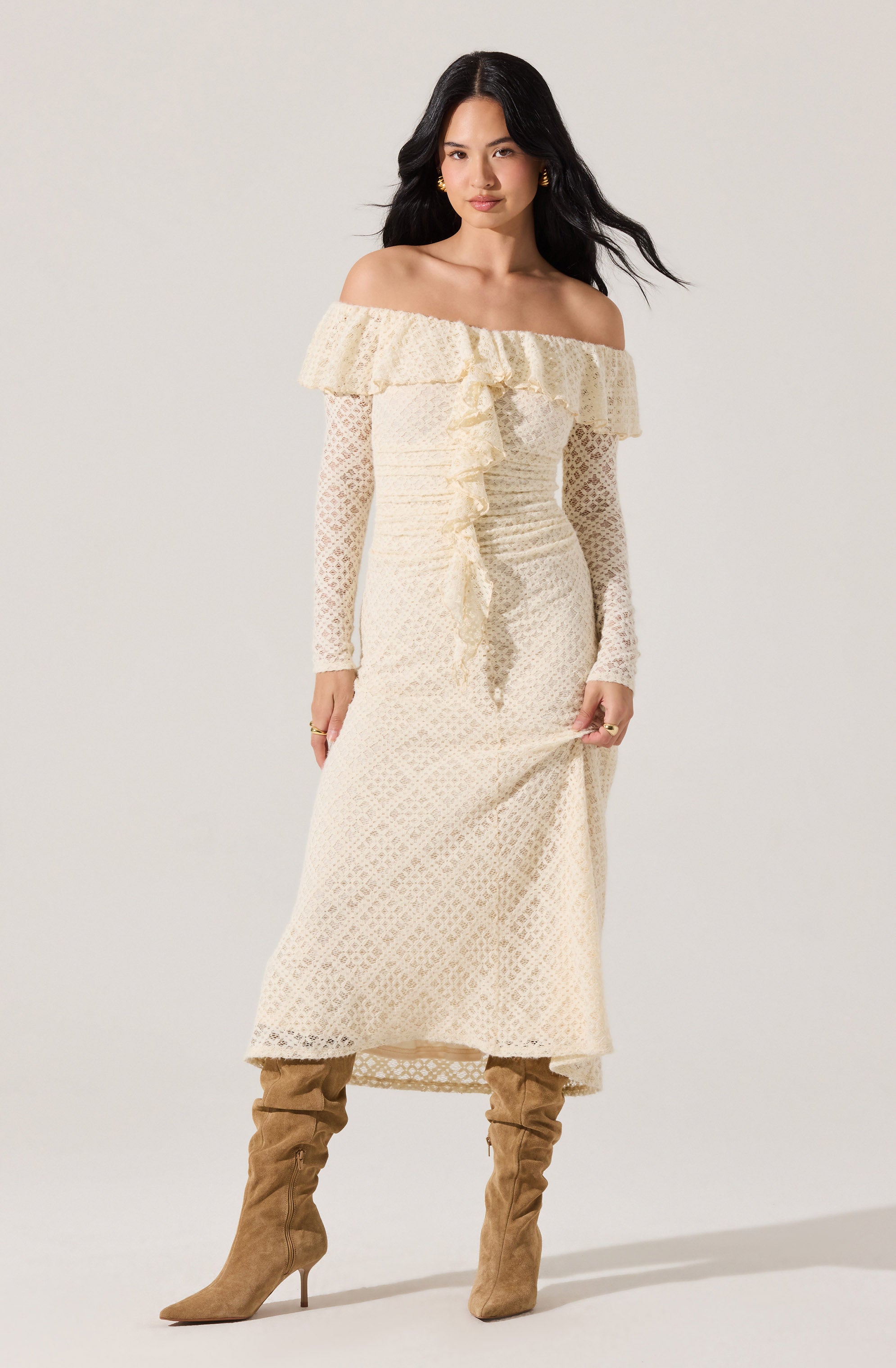 The woman is wearing a long, off-the-shoulder, cream-colored dress with a ruffled neckline and long, semi-sheer lace sleeves. She pairs the dress with knee-high, light brown suede boots with a pointed toe and stiletto heel.