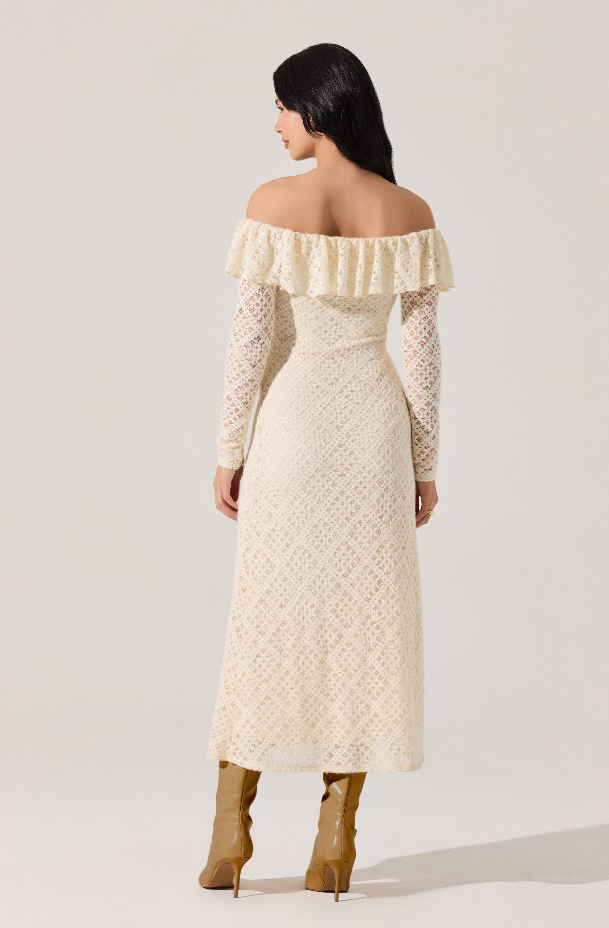 A woman is wearing an off-the-shoulder, long-sleeved dress in a light, cream color with a lace pattern. The dress features a ruffled neckline that drapes over the shoulders. She is also wearing tan knee-high boots with high heels.