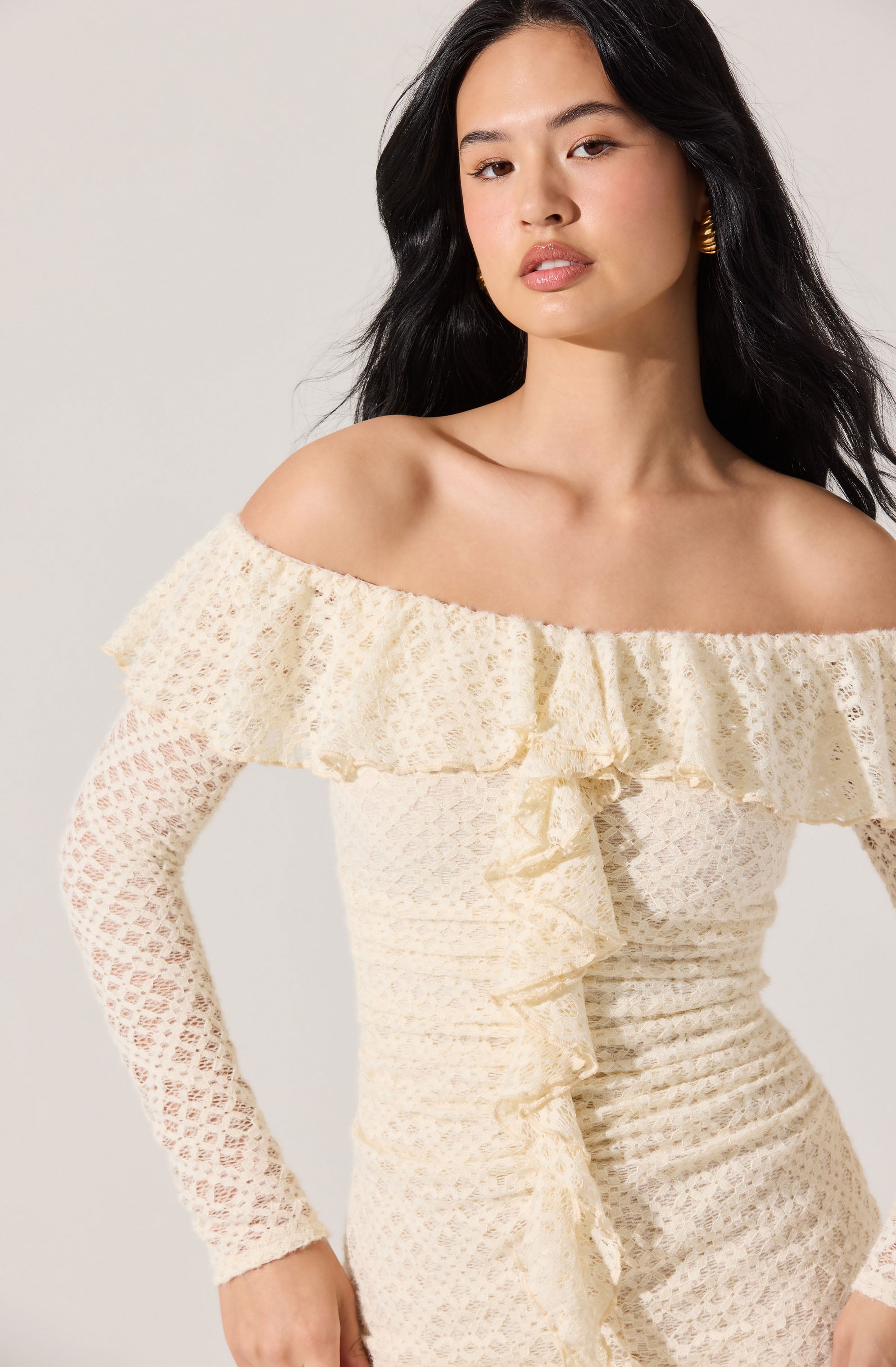 A woman is wearing an off-the-shoulder dress made from textured, light-colored lace fabric. The dress features a large ruffle detail along the neckline that extends around the shoulders and drapes down the front with a ruffled slit. The dress has long lace sleeves with a delicate, openwork pattern. She is accessorizing with large gold hoop earrings.