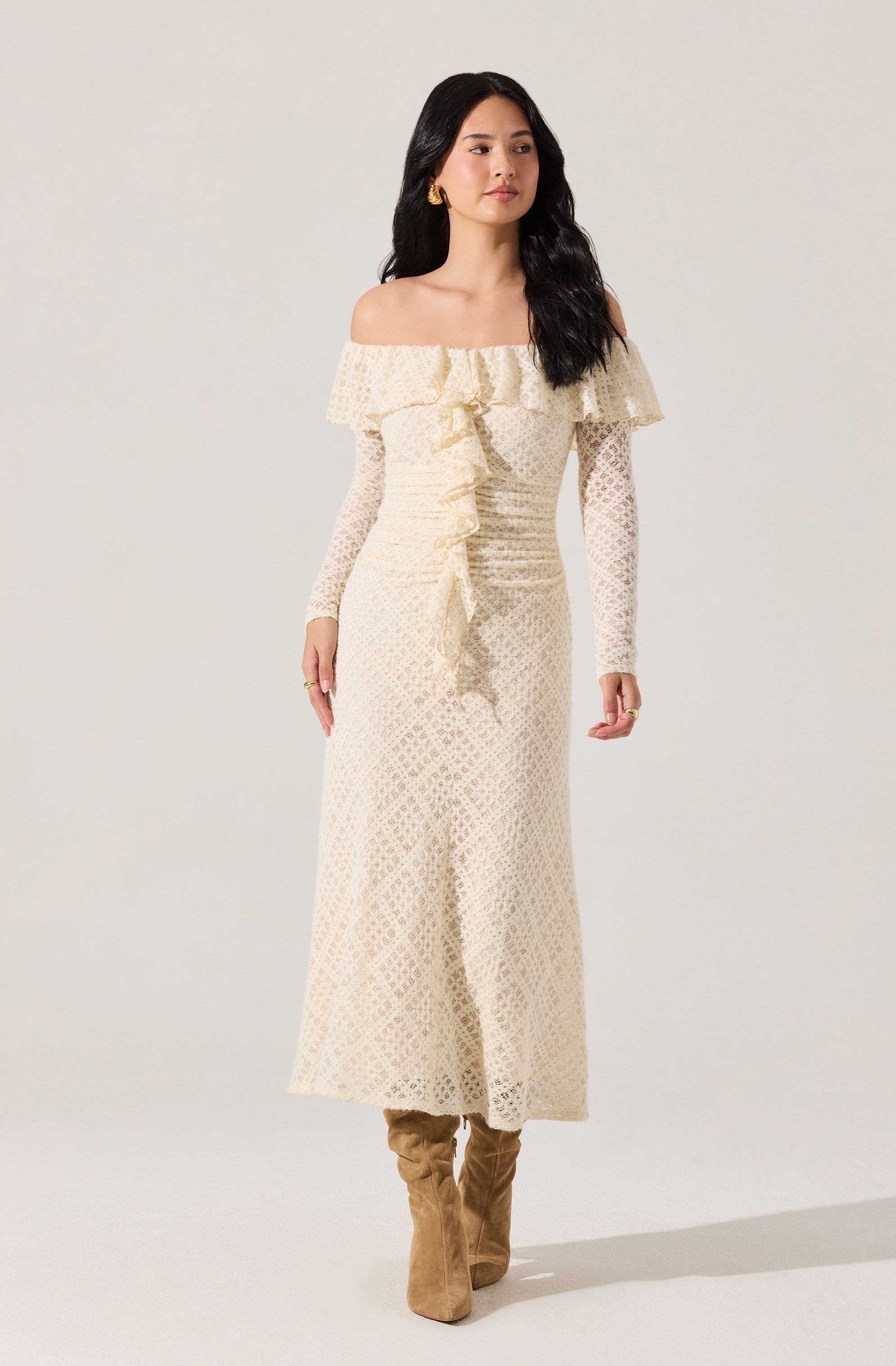 A woman is wearing an off-the-shoulder, long-sleeved cream-colored dress with a textured, lace-like pattern. The dress features a ruffled neckline that drapes over the shoulders and a ruffled front detail down the center. She is also wearing tan knee-high boots.
