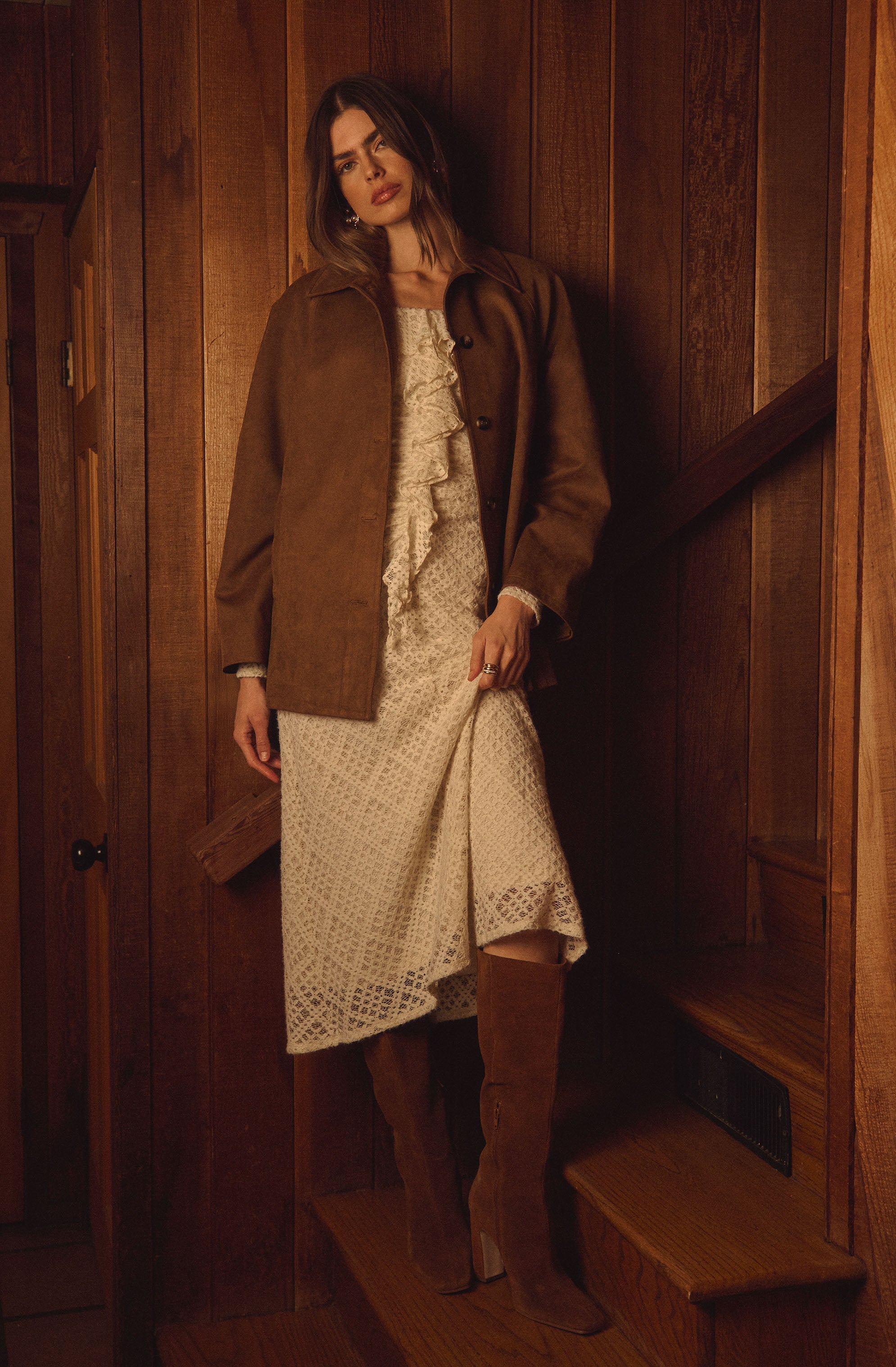 A woman is wearing a mid-length, cream-colored lace dress with ruffle details along the front and lacy cuffs at the sleeves. Over the dress, she has on a brown, button-up, collarless coat. She is also wearing tall, brown, suede high-heeled boots. The setting features warm, wooden paneling in the background.