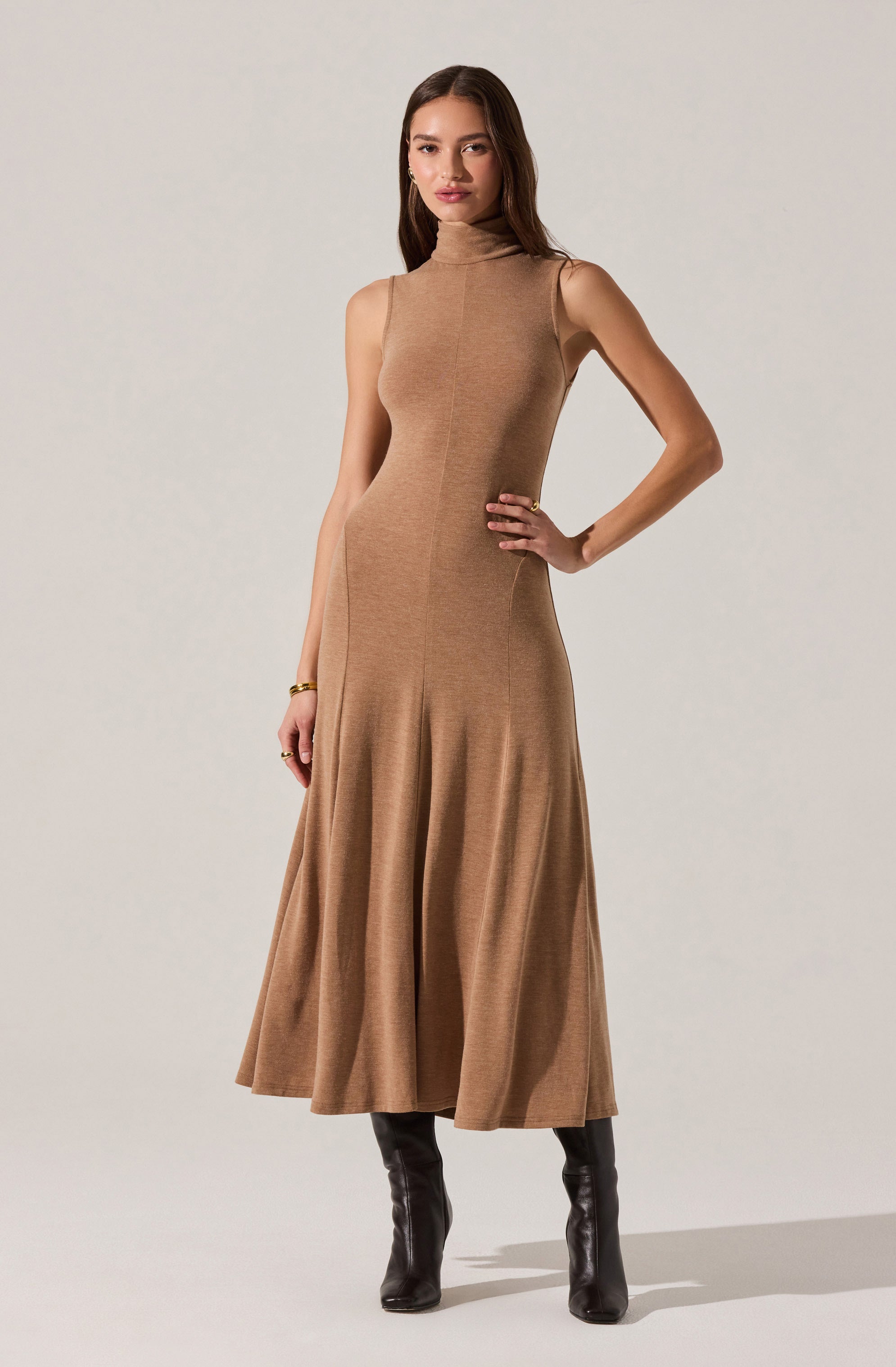 A woman is wearing a sleeveless, high-neck, camel-colored knit dress that is fitted through the bodice and flares out into a full-length skirt. She has paired the dress with black knee-high boots and is accessorizing with gold jewelry, including a bracelet and rings.
