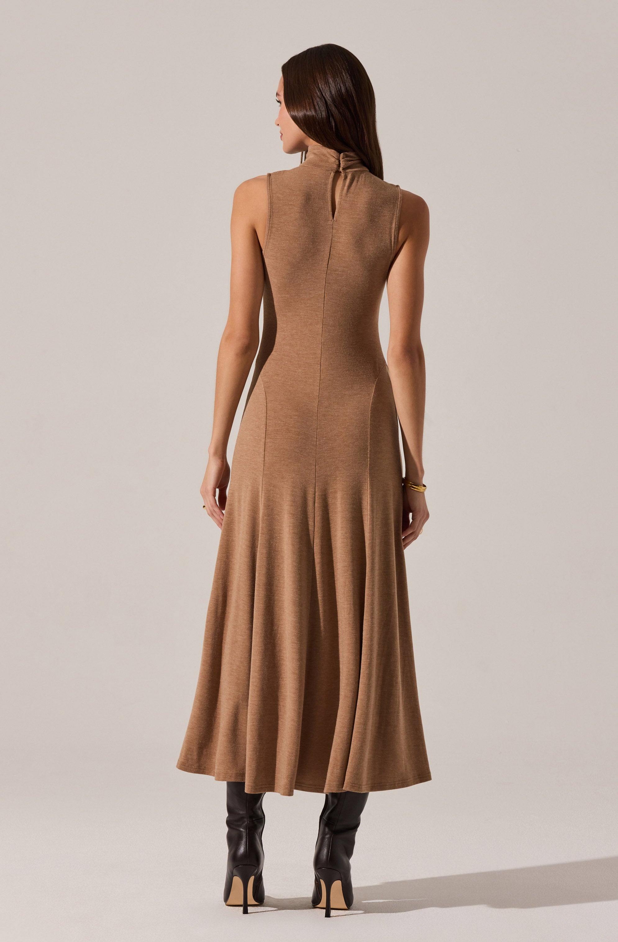 The woman is wearing a sleeveless, high-neck, camel-colored dress made from a textured fabric. The dress is fitted at the waist and flares out into a long, flowing skirt that reaches the floor. She is also wearing black high-heeled boots.