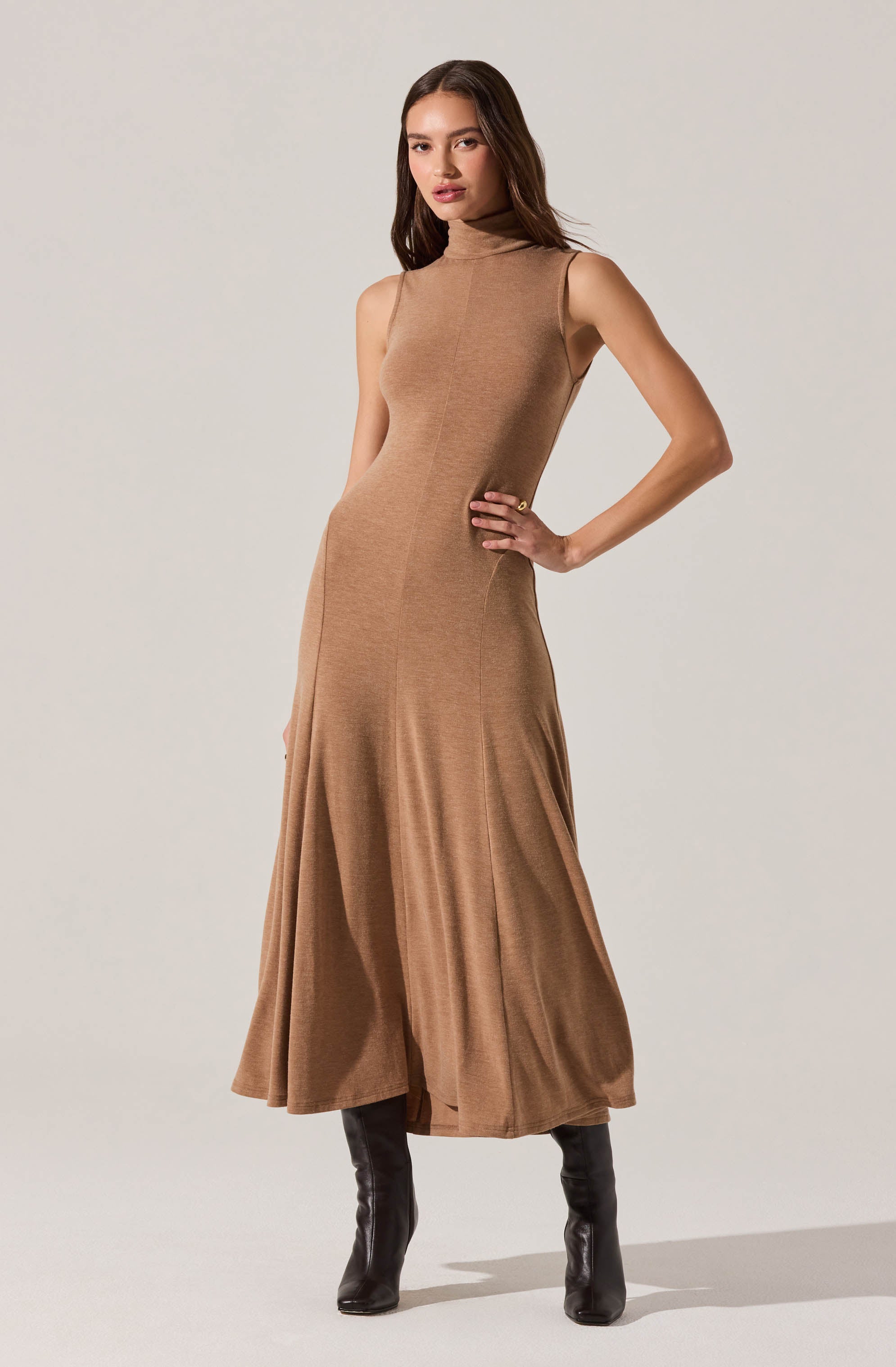 A woman is wearing a sleeveless, high-neck, camel-colored knit dress that is fitted through the waist and flows into a full, floor-length skirt with a slight flare. She pairs the dress with tall black boots.