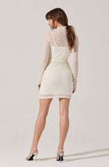 A woman is wearing a white lace dress with long sheer sleeves and a high neckline. The dress is form-fitting and short, ending above the knees. She is also wearing white high-heeled sandals with thin straps.