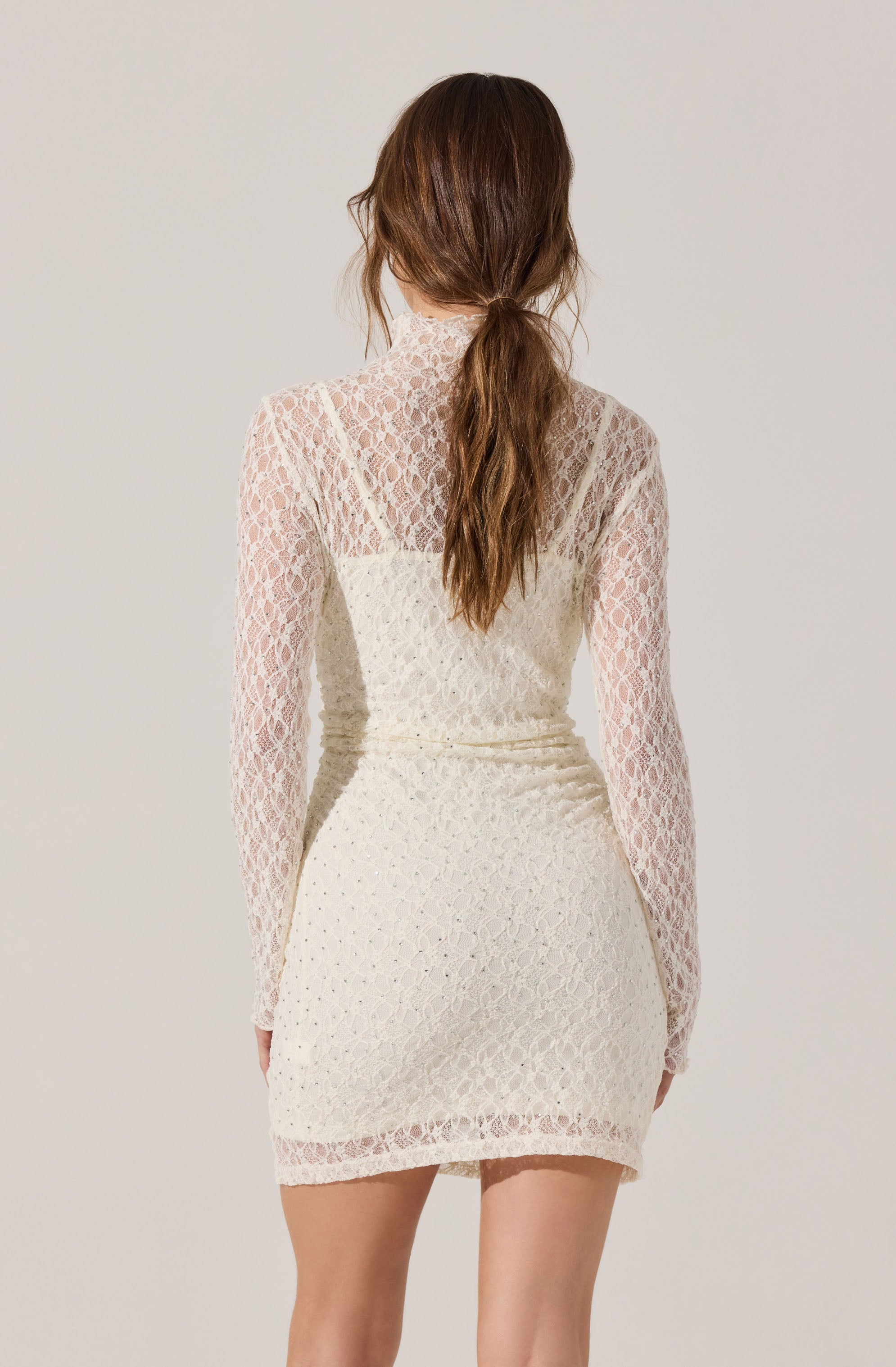 A woman is wearing a long-sleeved, high-neck dress made of white lace with a floral pattern. The dress has a fitted silhouette and falls to mid-thigh length. The lace fabric is see-through on the sleeves and upper back, with a lining on the rest of the dress. The woman’s long, wavy brown hair is styled in a loose ponytail.