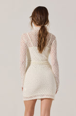 A woman is wearing a long-sleeved, high-neck dress made of white lace with a floral pattern. The dress has a fitted silhouette and falls to mid-thigh length. The lace fabric is see-through on the sleeves and upper back, with a lining on the rest of the dress. The woman’s long, wavy brown hair is styled in a loose ponytail.