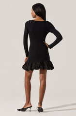 A woman is wearing a long-sleeved, black, ribbed knit dress that fits snugly and extends to just above her knees. The hem of the dress features a wide, ruffled, black fabric trim. She is also wearing black, pointed-toe high heels with white polka dots and a small bow detail on the side.