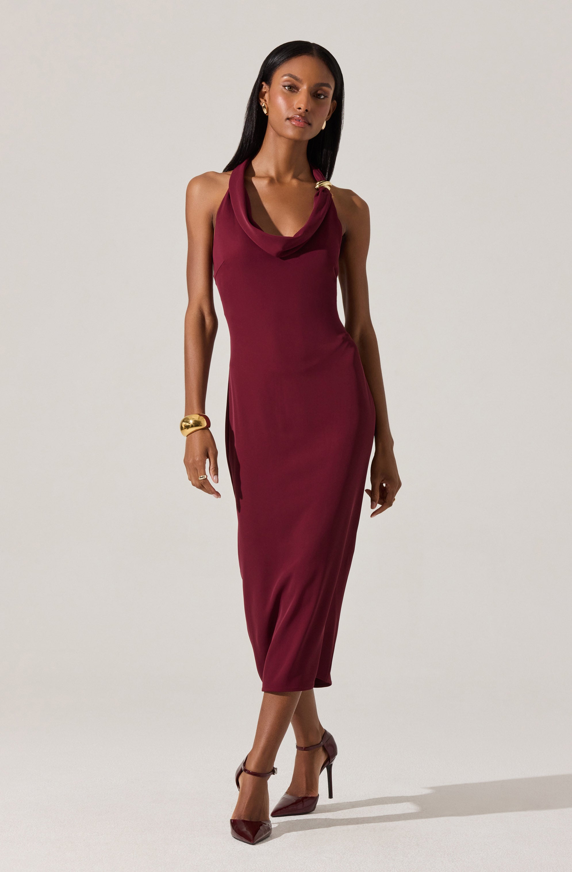 A woman is wearing a sleeveless, ankle-length burgundy dress with a draped neckline. The dress fits closely to the body, emphasizing her shape. She accessorizes with gold jewelry, including a large bracelet on her right wrist and earrings. She is also wearing high-heeled, pointed-toe shoes that match the color of her dress.
