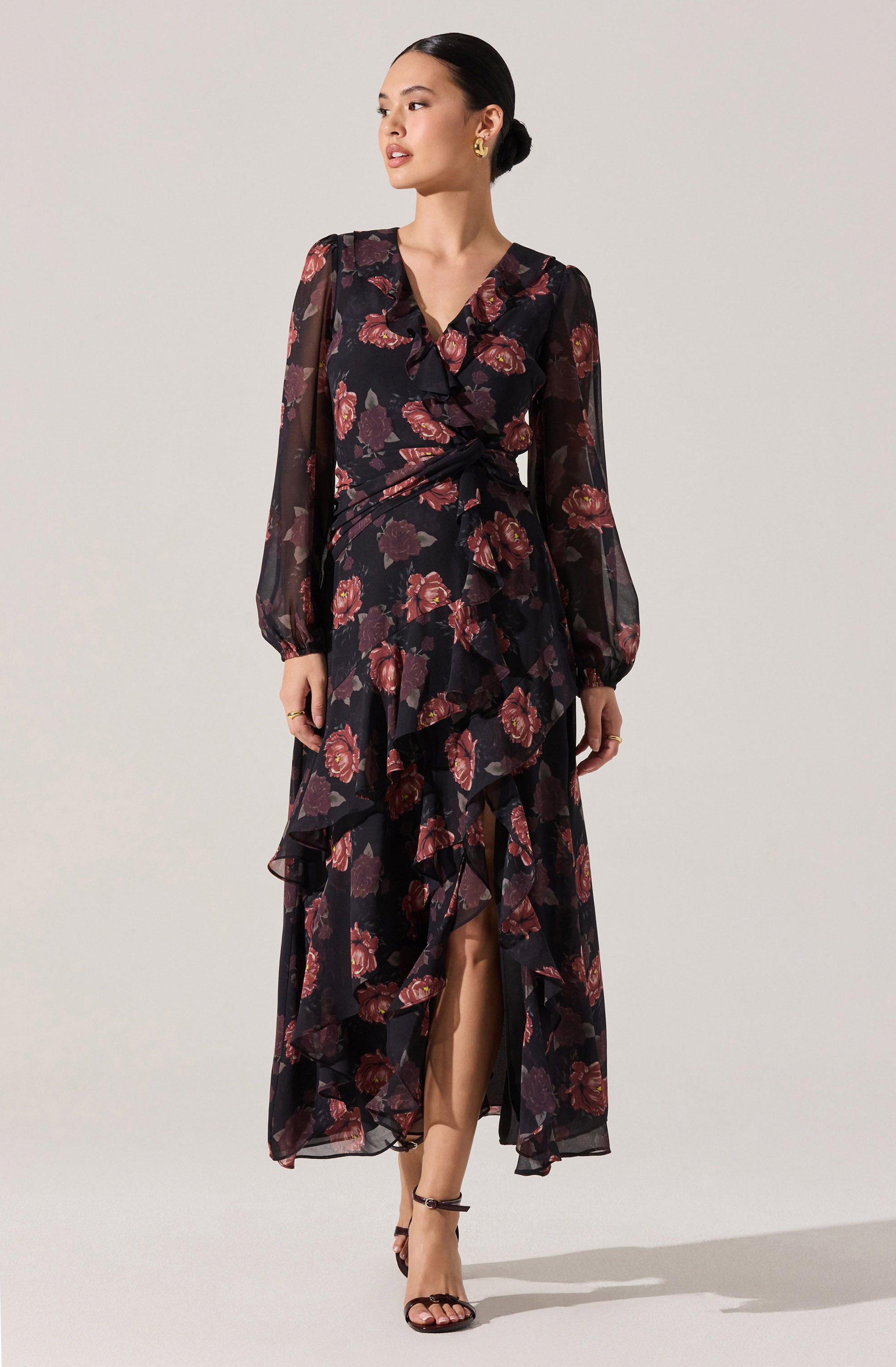 A woman is wearing a long, flowy black dress with a floral pattern featuring pink and purple flowers. The dress has long, sheer sleeves and a ruffled, layered skirt with a slit in the front. The neckline is decorated with a ruffle trim.