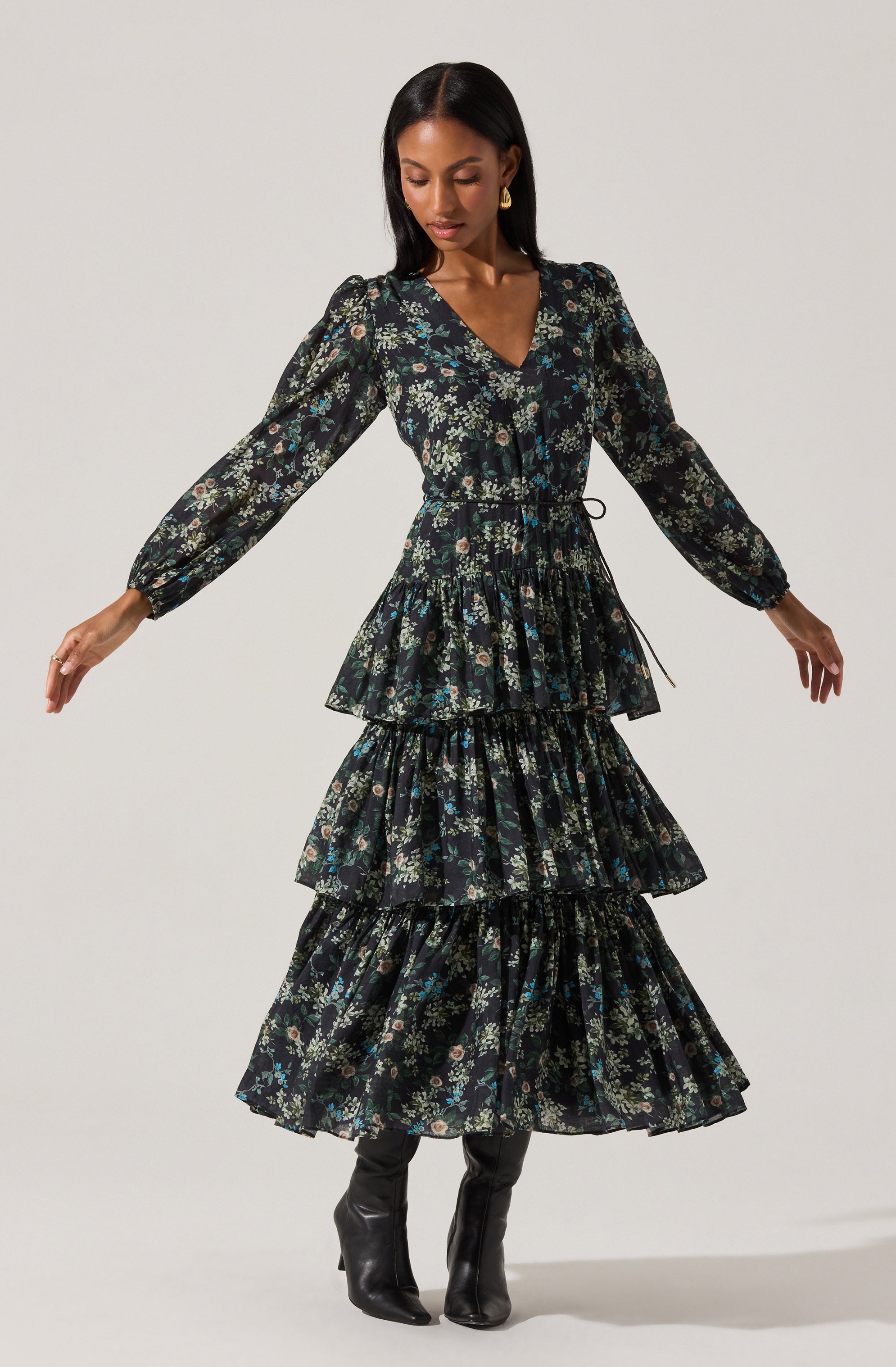 A woman is wearing a long, dark floral dress with a V-neckline and long, puffed sleeves. The dress features a fitted waist and three tiered, ruffled skirt sections. She pairs the dress with black knee-high boots and gold earrings.