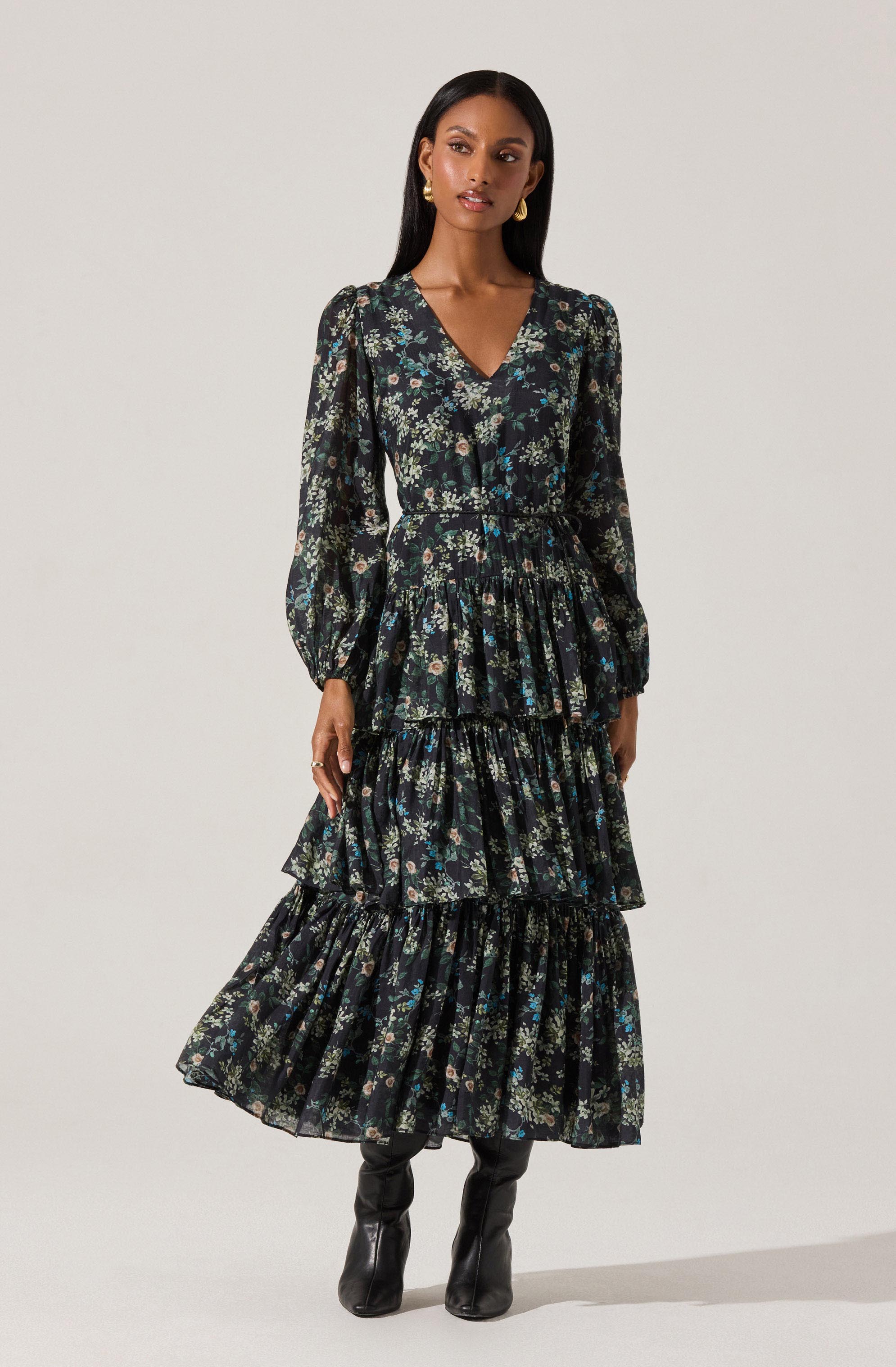 A woman is wearing a long, black floral dress with a V-neckline. The dress features a tiered skirt with multiple ruffles and long, slightly puffed sleeves. The floral pattern includes small flowers in shades of blue, green, and beige. She completes her outfit with black knee-high boots and gold earrings.