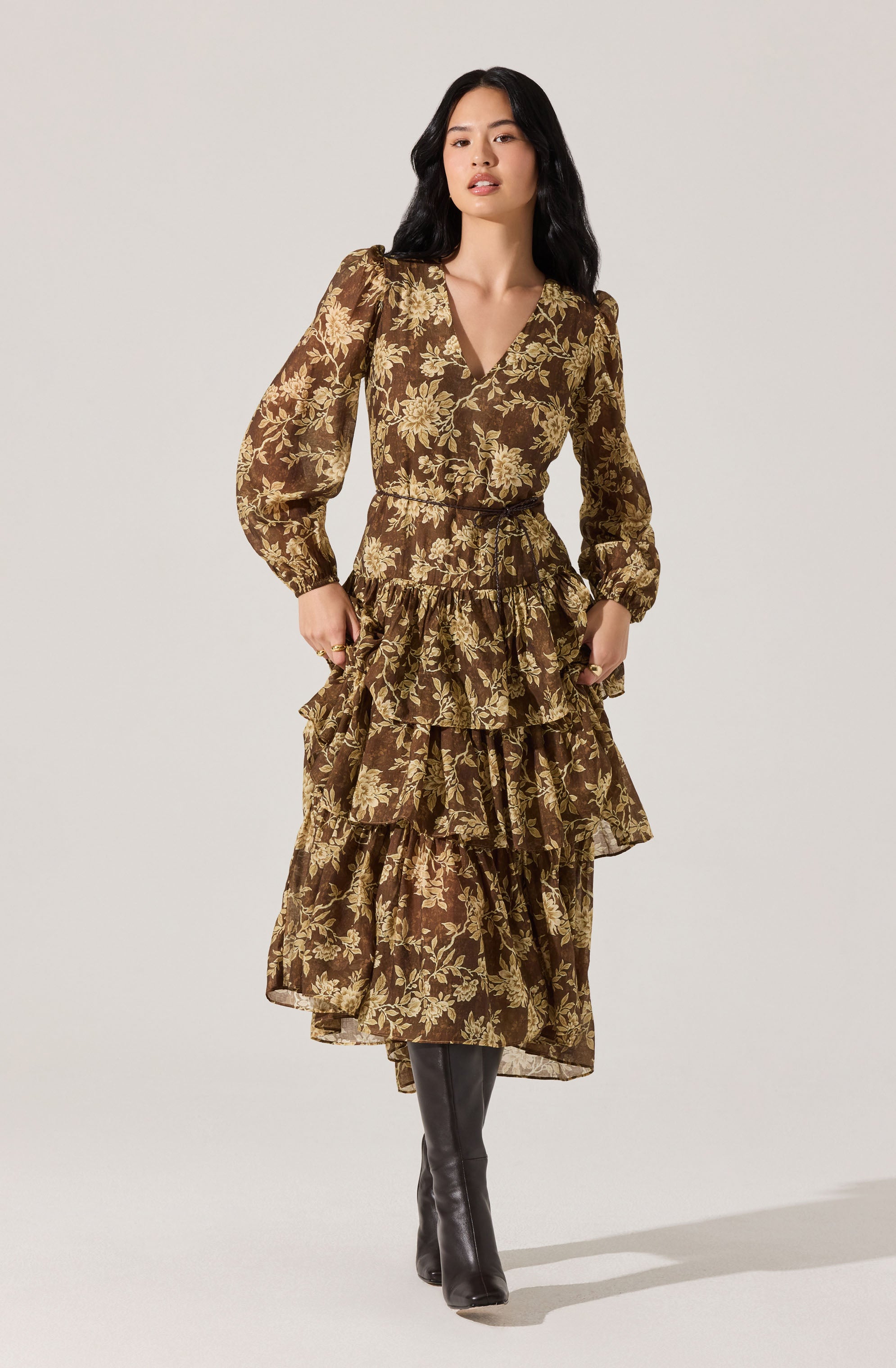 A woman is wearing a long, brown dress with a floral pattern in shades of beige. The dress has a V-neckline, long puffed sleeves, a fitted waist, and multiple tiered ruffles that extend down to her ankles. She is also wearing black knee-high boots.