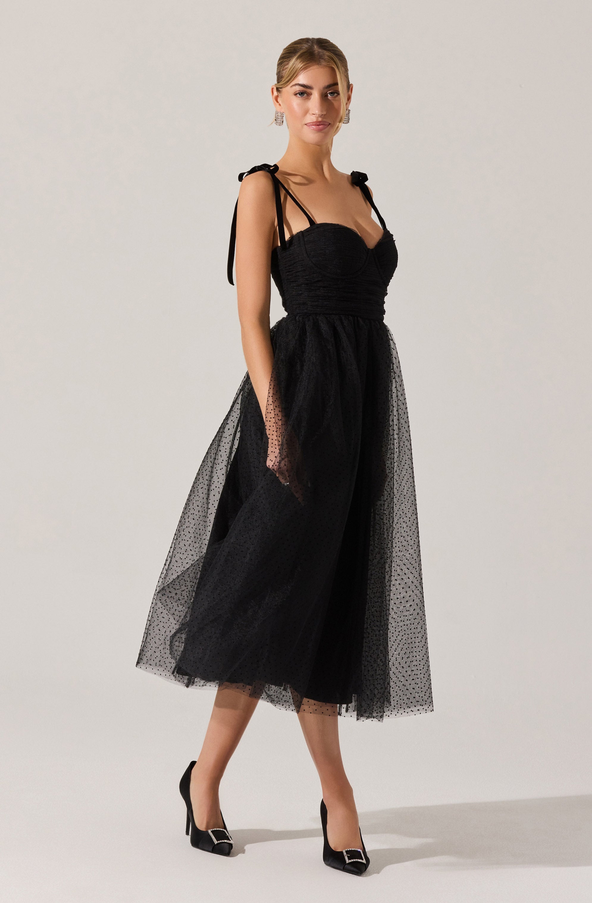 A woman is wearing a black dress with a fitted, ruched bodice and thin shoulder straps tied into bows. The skirt is made of a sheer, polka-dotted tulle fabric, creating a layered, voluminous look that reaches mid-calf. She pairs the dress with black high-heeled shoes featuring pointed toes and metallic embellishments on the front.