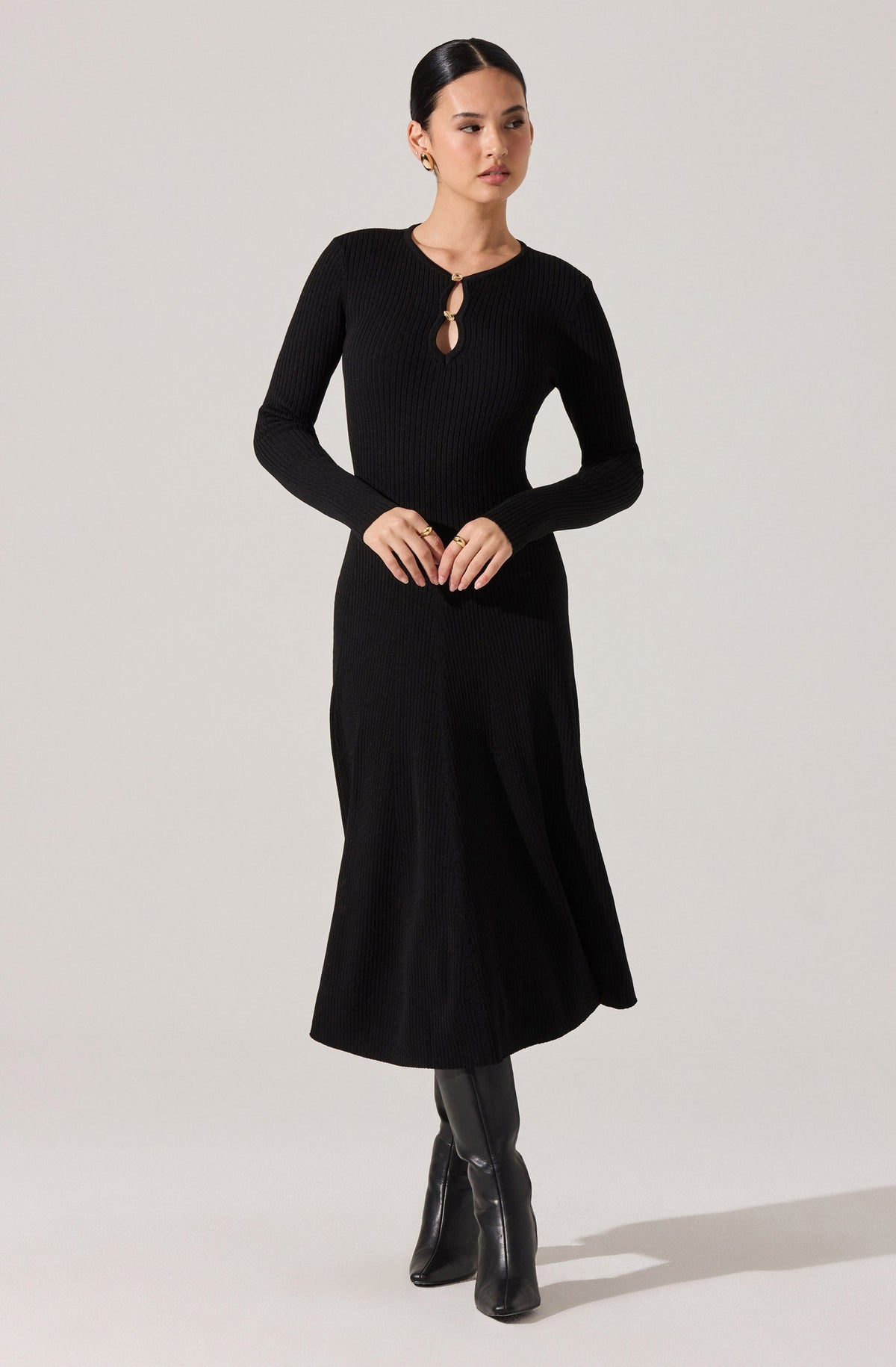 A woman is wearing a long black ribbed knit dress with long sleeves. The dress features a small keyhole opening at the neckline, secured with a round button. She completes the look with black knee-high boots.