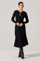A woman is wearing a long black ribbed knit dress with long sleeves. The dress features a small keyhole opening at the neckline, secured with a round button. She completes the look with black knee-high boots.