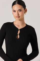 A woman with dark, straight hair styled in a sleek bun is wearing a black, ribbed, long-sleeve top with a round neckline. The top features a small keyhole opening near the collar, fastened with two beige, shell-shaped buttons. She has gold hoop earrings.