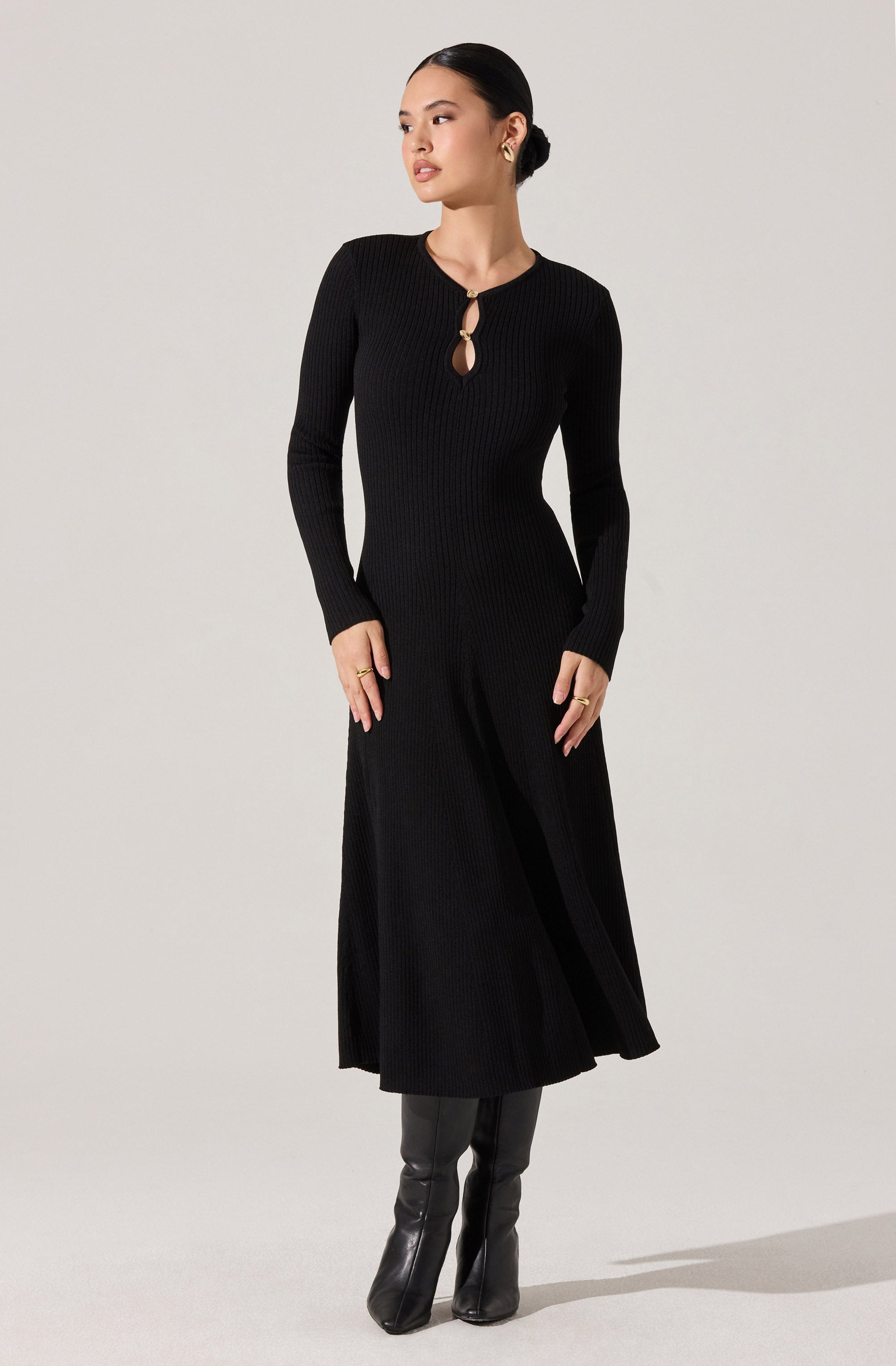 A woman is wearing a long, black, ribbed knit dress with a round neckline and a small keyhole opening secured by a circular knot detail. The dress has long sleeves and a fitted waist, flowing into an A-line skirt that reaches her ankles. She is also wearing black knee-high boots.