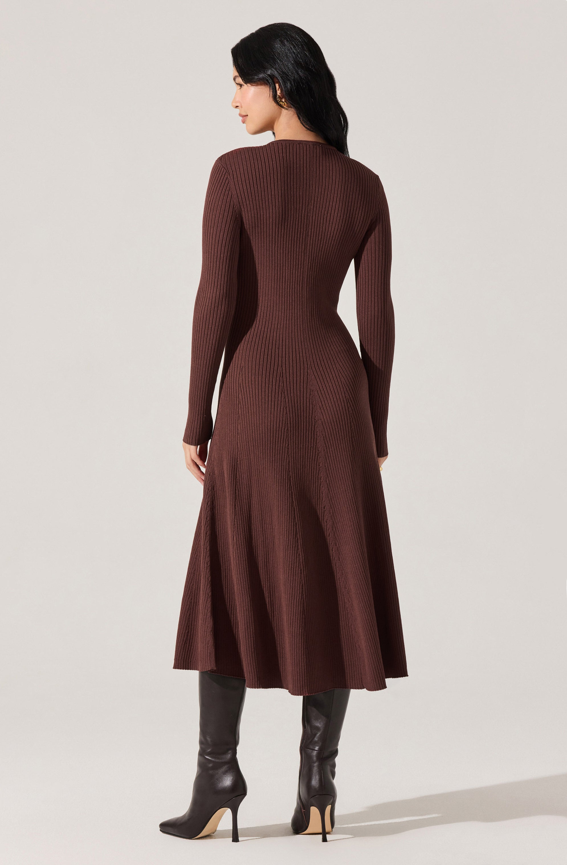 A woman is wearing a long, ribbed, chocolate brown dress with long sleeves and a fitted waist that flares into a flowing skirt. She is also wearing tall, pointed-toe black high-heeled boots.