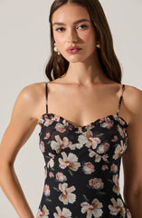 A woman is wearing a black sleeveless dress with thin spaghetti straps. The dress features a floral pattern of large, light-colored flowers with pink and beige petals and green leaves. The top of the dress has a ruffled detail along the neckline. She is accessorized with large, statement earrings that have a pearl and gold design.